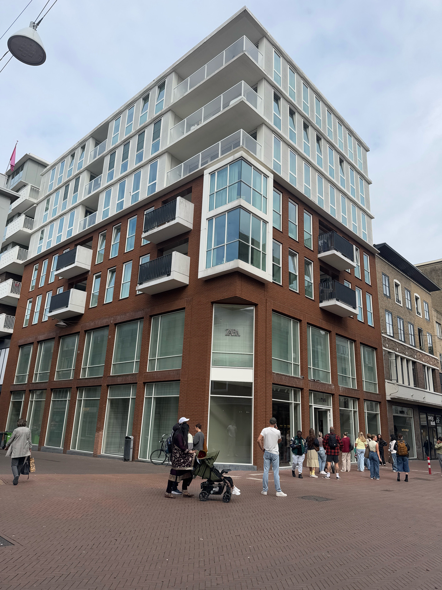 A low-angle, wide shot shows a modern, multi-story building with red brick lower floors and white upper floors with balconies. The building's facade has large windows and balconies. On the ground floor, the "ZARA" logo is visible on a glass section of the building. A group of people are walking on the brick-paved sidewalk in front of the building. Some individuals are looking into the store windows. The sky is overcast. The scene appears to be a commercial street in Nijmegen, Netherlands.