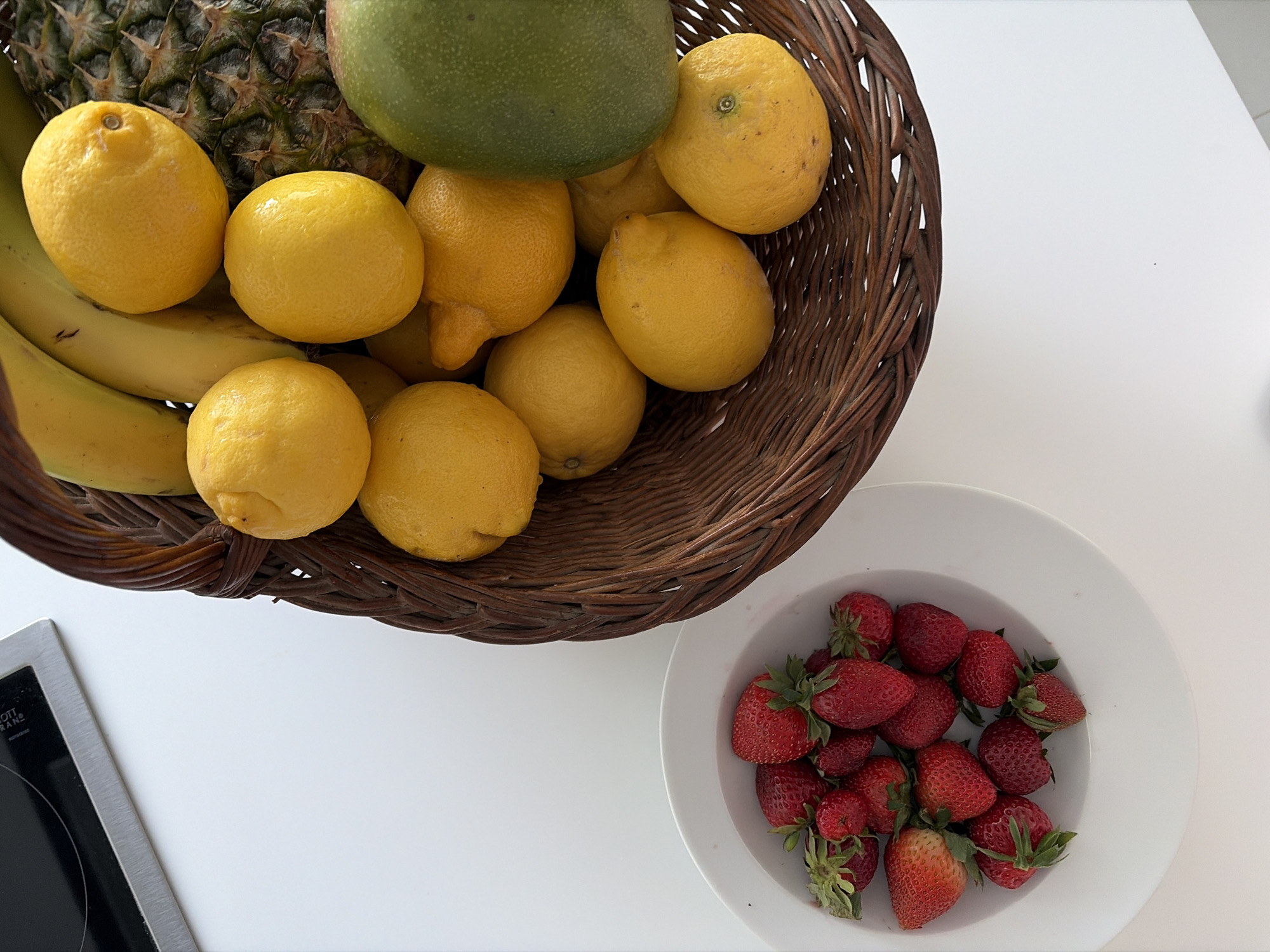 A high-angle shot depicts a white tabletop with a wicker basket filled with fruit and a white bowl of strawberries. The basket contains bananas, lemons, a pineapple, and a green mango. The bowl of strawberries is positioned to the right of the basket. A portion of a built-in stovetop is visible in the lower left corner. The image does not provide enough information to determine a specific location within the Municipal Unit of Elliniko, Greece.
