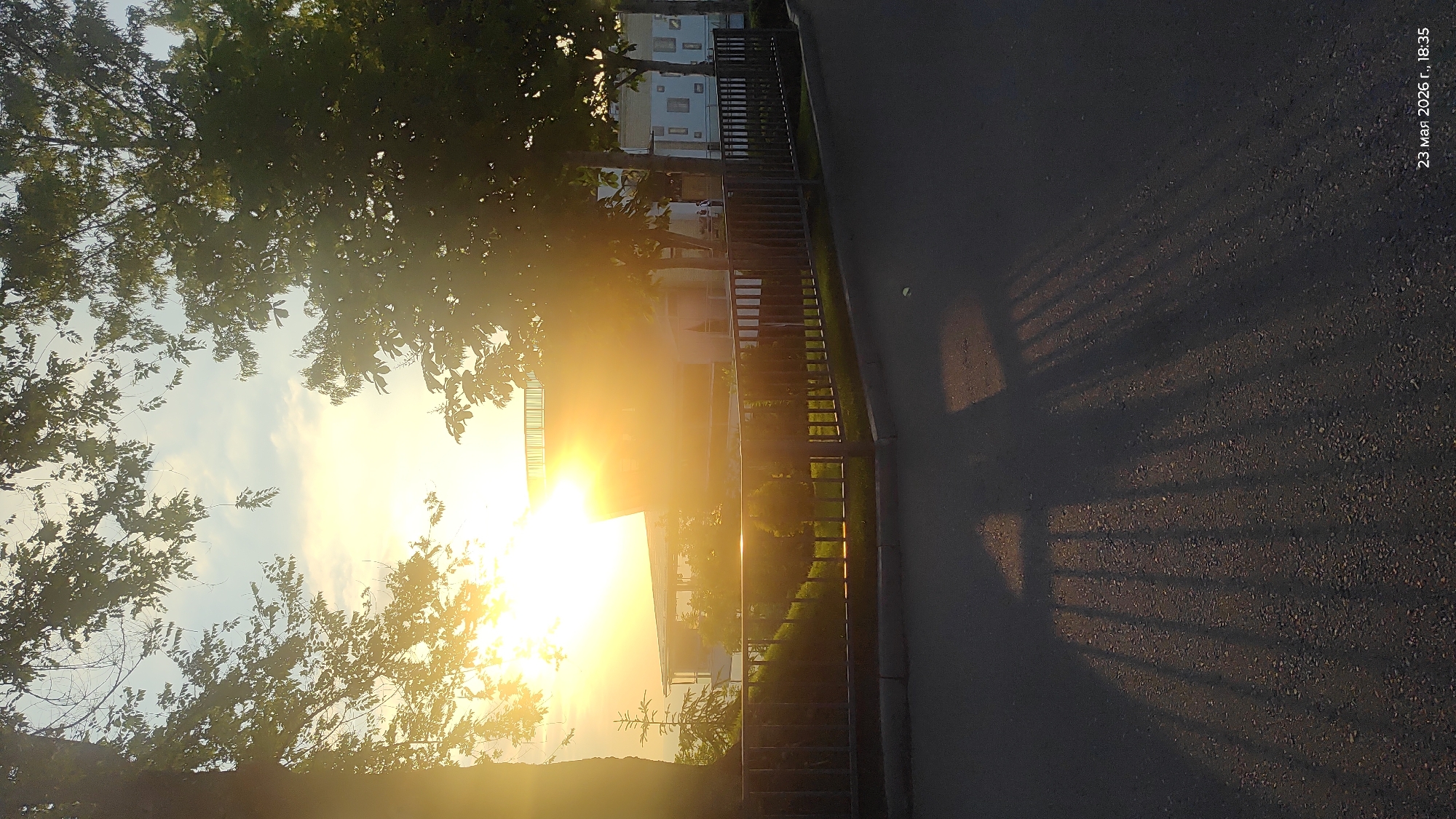 The image shows a sunset scene in what is likely a residential area. The sun is low on the horizon, creating a strong glare. In the foreground is a sidewalk and a metal fence that casts a shadow on the pavement. The setting appears to be urban, with a building visible behind the fence and sunlight. Trees with dense foliage are situated on the left side of the frame, with the setting sun visible through the branches. On the right side of the frame, there is also the date "23 March 2026 r., 18:35". Without additional information, pinpointing the location in Almaty is not possible.