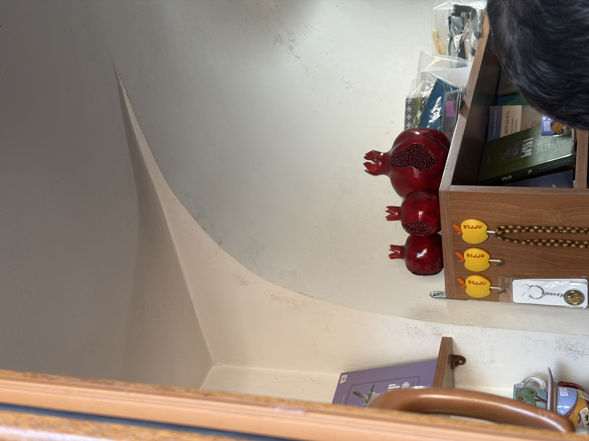 This is a close-up, low-angle, interior shot of a shelf and wall in Ashtarak, Armenia. The scene focuses on a collection of decorative items and a portion of a person's head. The wall behind the shelf has a curved archway.
On the shelf, there are three red decorative pomegranates. The largest one in the center is carved to appear partially open, revealing its seed-like interior. To the left and right of it are two smaller, solid red pomegranates. Behind these, on the shelf, are several items wrapped in plastic, appearing to be fabrics or books.
Below the shelf, on a wooden board, are three yellow, apple-shaped magnets. Hanging from hooks beneath the apples are a keychain with a circular pendant and a string of brown beads.
To the left of the shelf, a dark wooden frame or doorjamb is visible. On a lower shelf, a ceramic mug with a painted tree design is partially visible, along with other patterned ceramic items.
A person's head, with dark, curly hair, is visible in the lower right corner of the image, looking downwards towards the shelf.