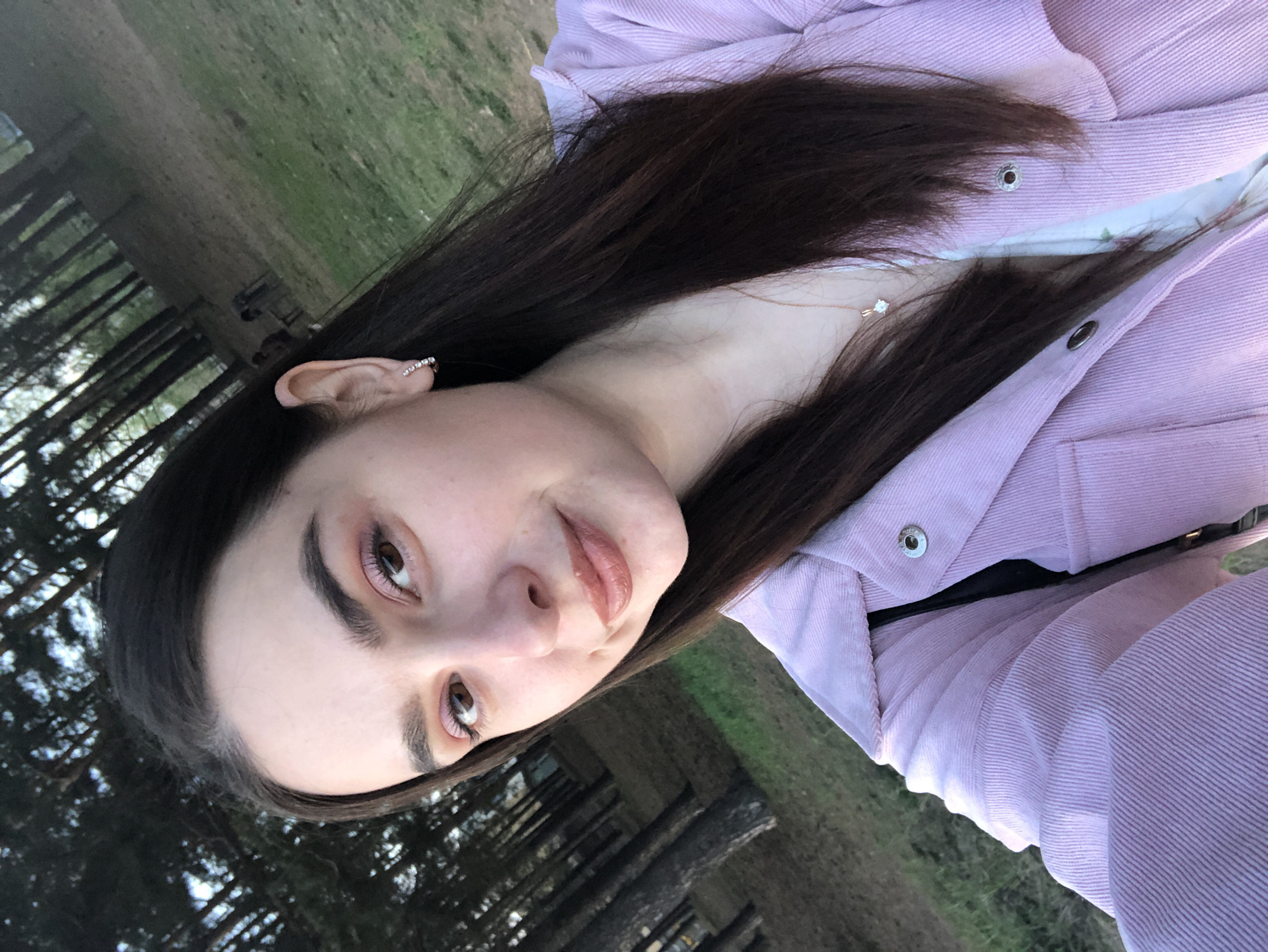 A young woman poses in an outdoor setting, potentially a park or wooded area near the city of Chaikovsky, Russia. The background consists of trees and grassy terrain, suggesting a natural environment. The woman is wearing a lavender corduroy jacket over a white top and a delicate gold necklace. She has long, dark hair and light brown eyes. She has earrings in her left ear. The lighting indicates a daytime setting.