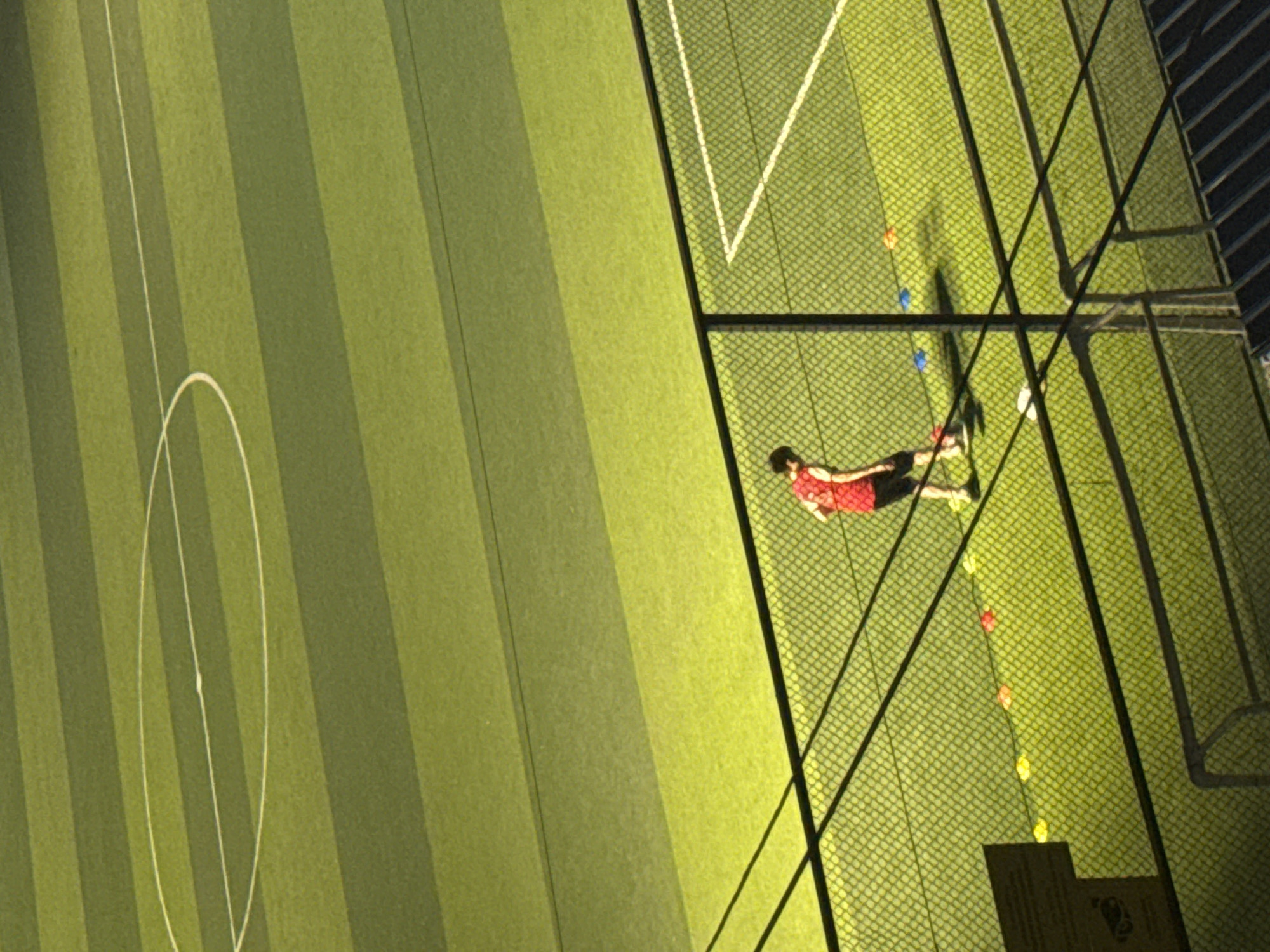 An elevated, full shot captures a person in athletic attire on a brightly lit, artificial turf soccer field in Tbilisi, Georgia. The field is marked with white lines, including a half-circle on the upper portion of the image. The person, wearing a red jersey and black shorts, is positioned in the mid-ground, performing some sort of drill with brightly colored cones of red, yellow, and blue scattered on the field near them. A chain-link fence runs across the foreground, partially obscuring the view of the soccer field and the person. The lighting suggests it is nighttime or dusk.