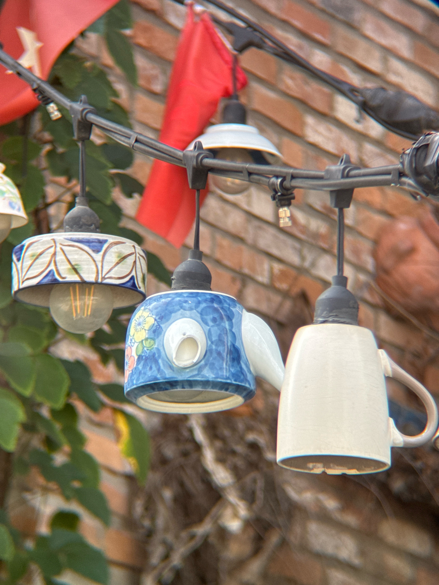 In Nha Trang, Vietnam, an outdoor scene features a string of lights suspended above a brick wall. The lights are unique, with some housed in decorative ceramic shades resembling pottery, one appearing to be a painted teapot, and another in a white mug. Green foliage is visible to the left, and a red flag, possibly part of a banner, hangs in the background. The overall impression is of a whimsical, creatively decorated outdoor space.
