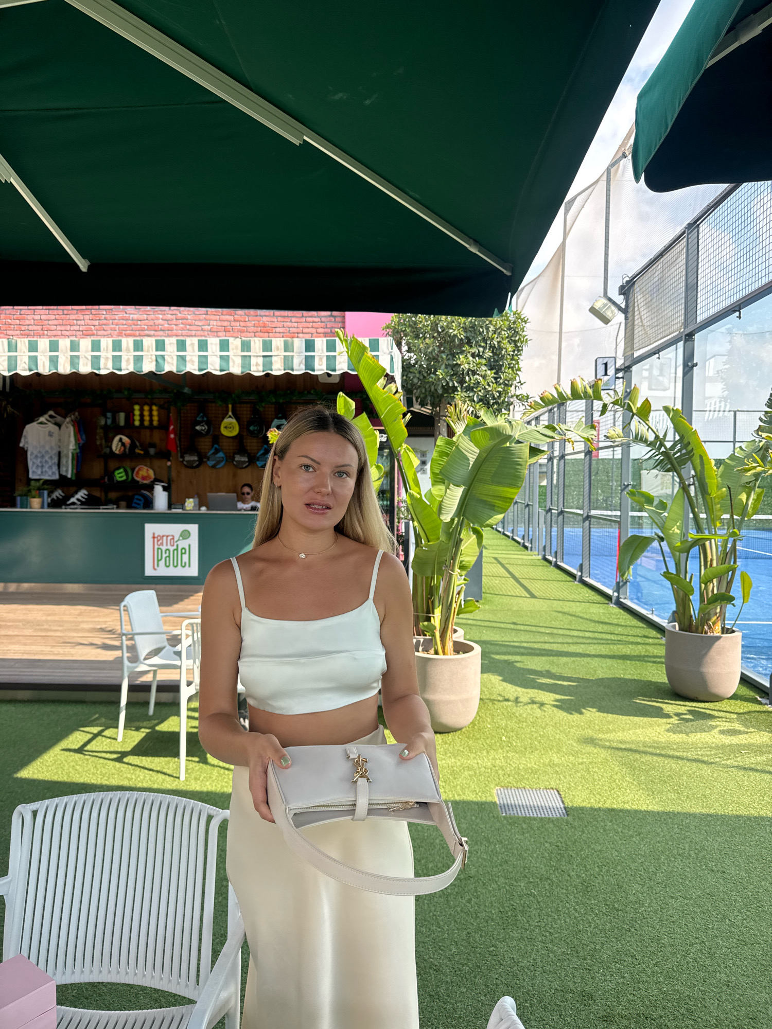 The image depicts a woman standing outdoors in what appears to be a sports facility, specifically a padel club, as indicated by the "terra padel" logo on the building in the background. The location is identified as Antalya, Turkey.
The woman, positioned in the foreground, is wearing a white satin crop top and a matching satin skirt. She is holding a light-colored handbag. Her blonde hair is down, and she is looking towards the camera with a neutral expression.
Behind her, a building with a brick facade and a green awning is visible. This building houses a counter with various sports equipment, including padel rackets and balls, suggesting it is a pro shop or reception area. White tables and chairs are arranged on a wooden deck in front of the building.
The environment surrounding the woman is characterized by artificial green turf, a blue padel court enclosed by a glass and metal fence, and large potted plants with broad leaves. The scene is illuminated by natural daylight, casting shadows from the plants and awnings. The overall atmosphere suggests a sunny day at a recreational facility.