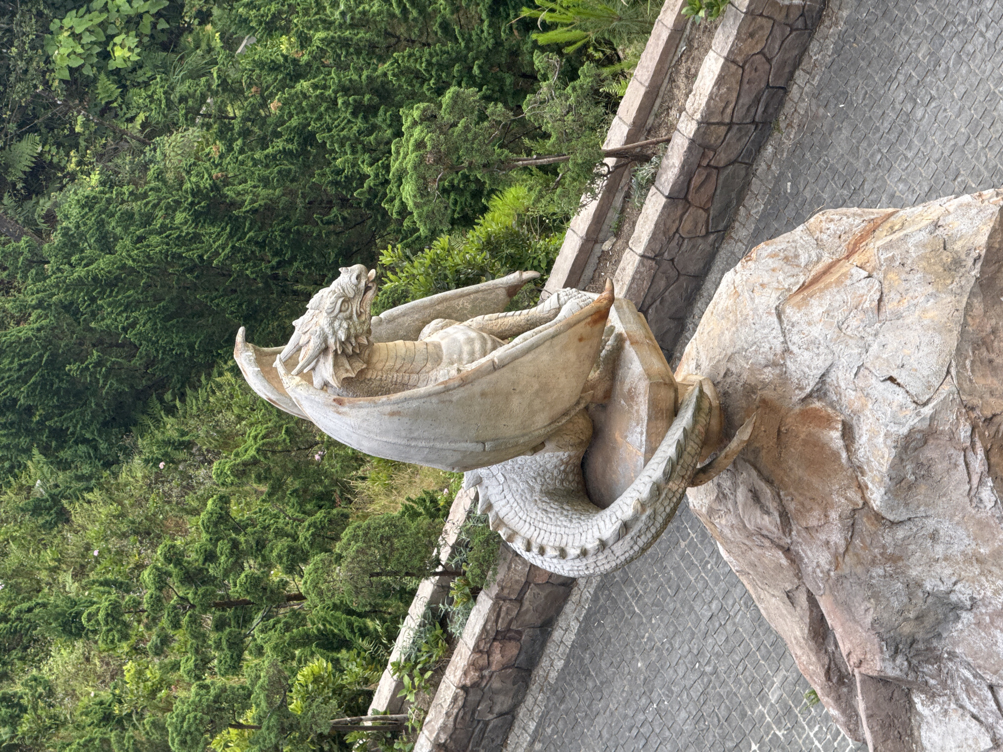 The image depicts a stone dragon statue in Đà Nẵng, Vietnam. The statue is positioned atop a stone structure, near a cobblestone path and lush green foliage. The dragon has a serpentine body, scaled texture, and a detailed head. The environment suggests a park or garden setting.
