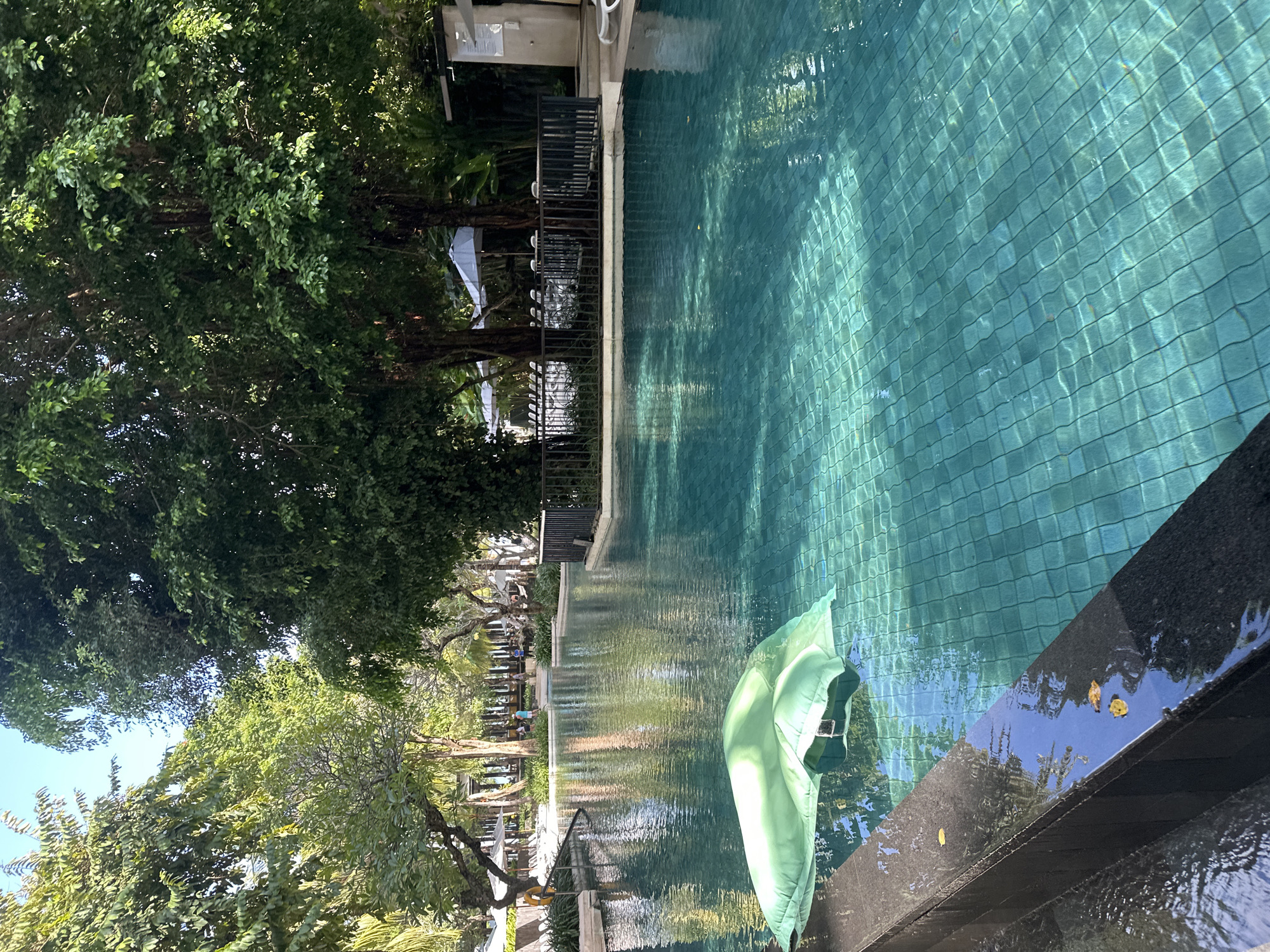 A wide shot captures a resort swimming pool with turquoise tiled water, reflecting sunlight and lush green trees. In the foreground, a deflated, light green inflatable rests on the pool's surface near the dark, polished stone edge. Beyond the pool, lounge chairs and umbrellas are visible, along with several people walking in the distance, suggesting the location is a resort in Kuta, Indonesia. The scene is peaceful, bathed in natural light.