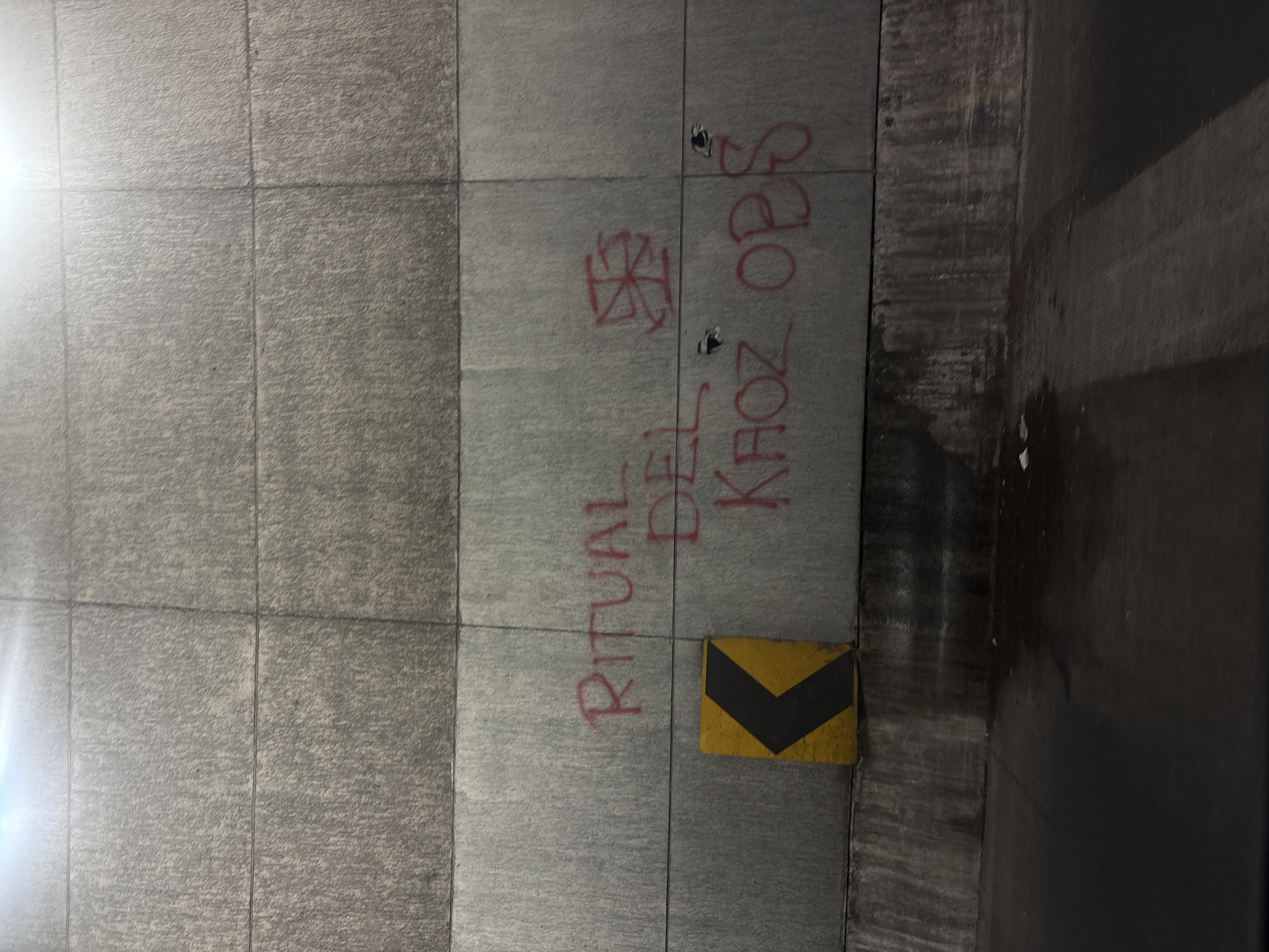 A photograph taken in Mexico City, Mexico, depicts a concrete wall with red graffiti. The wall is constructed from large rectangular blocks with visible seams. The graffiti reads "RITUAL DEL KHOZ OPS" and includes a swastika symbol. A yellow and black chevron marker is attached to the wall near the bottom. The floor is dark and wet, suggesting an indoor location like a parking garage or tunnel.