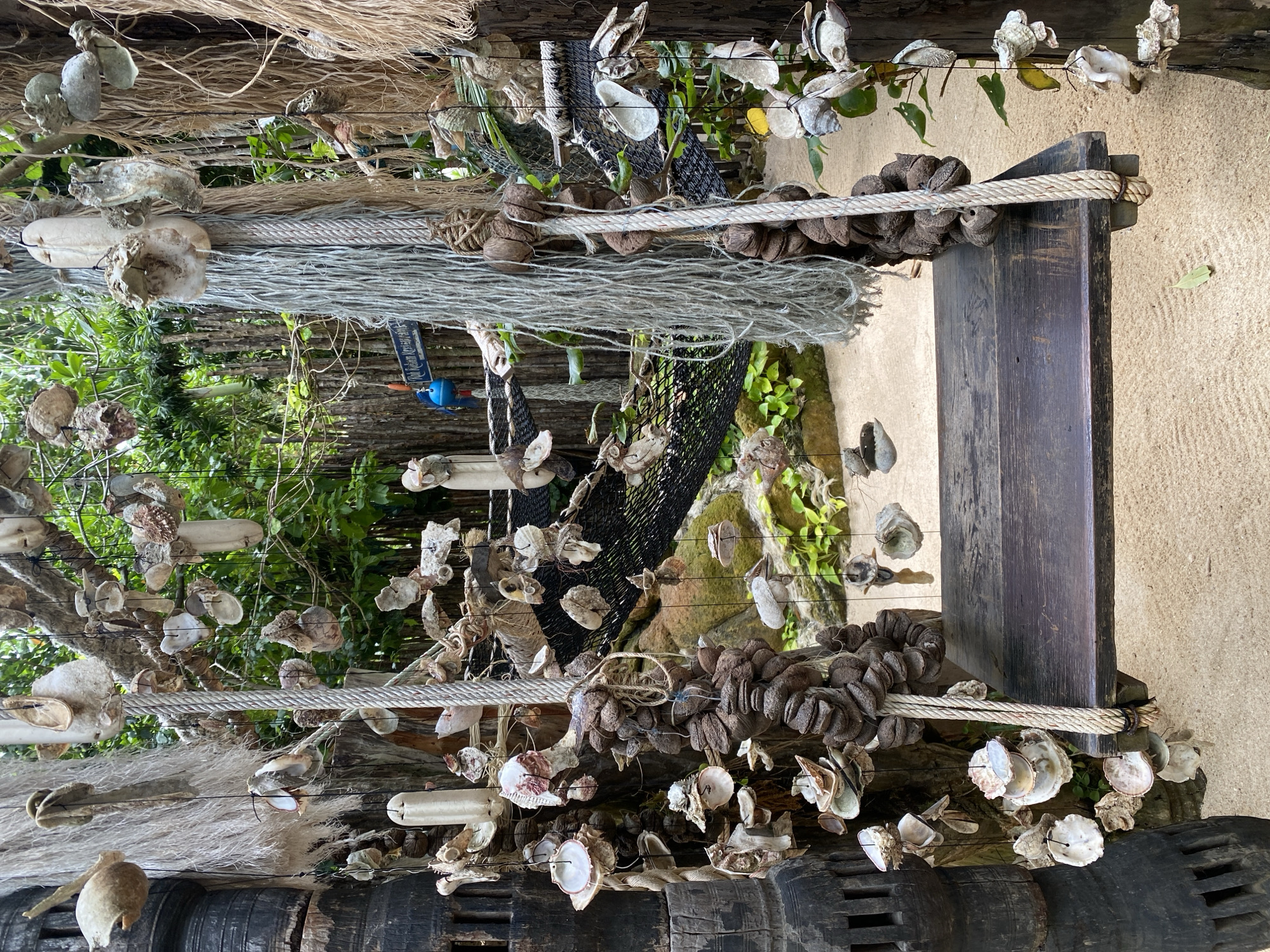 The image shows a rustic swing made of dark wood suspended by thick ropes. The swing is positioned on a sandy surface, possibly a beach or a garden path. The background is lush with greenery and a wooden fence, suggesting an outdoor tropical setting. The swing is adorned with numerous seashells and other natural objects, creating a unique and decorative aesthetic. To the left of the swing, a large, dark, carved wooden structure with vertical slots is visible. To the right, a thin rope with shells hanging from it extends downwards. In the middle ground, a black net is suspended, and further back, there are more natural elements and foliage. The overall scene evokes a sense of a coastal or island retreat. The location is identified as Ko Pha Ngan Subdistrict, Thailand.