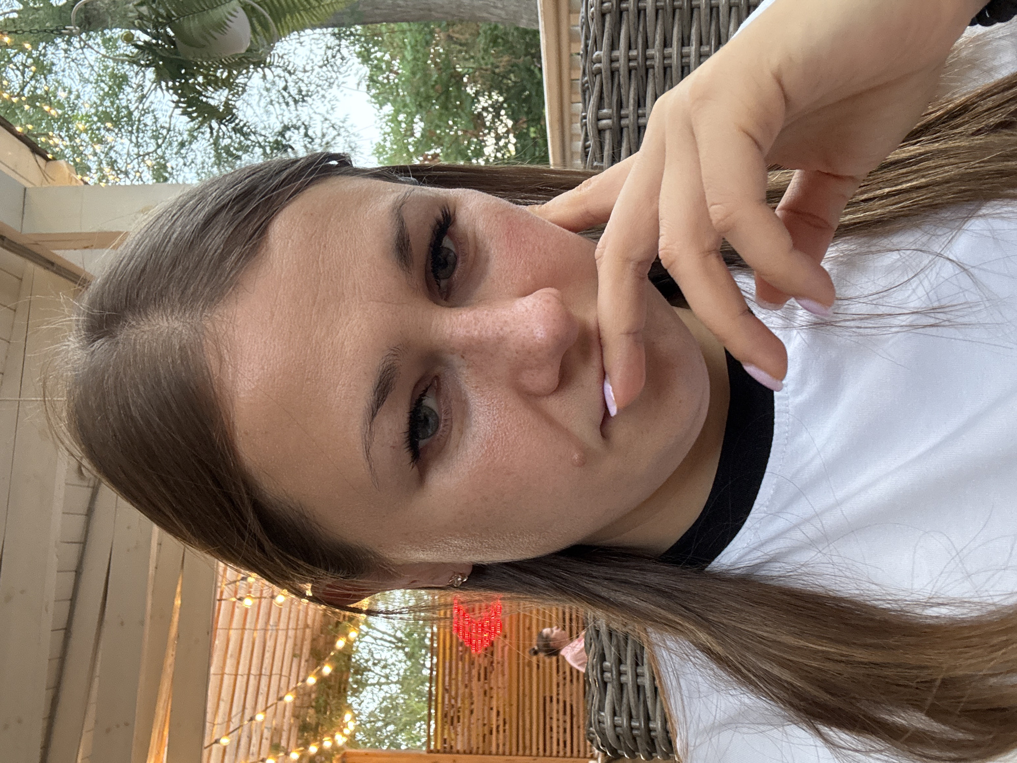 A woman is taking a selfie outdoors in Yevpatoriya, Ukraine, during daylight. She has long brown hair and is wearing a white t-shirt with a black collar. Her right hand is brought up to her mouth, with her fingers touching her lips. In the background, there is a wooden fence with string lights, and a person is seated at a table. The environment appears to be a patio or outdoor seating area with wicker furniture.