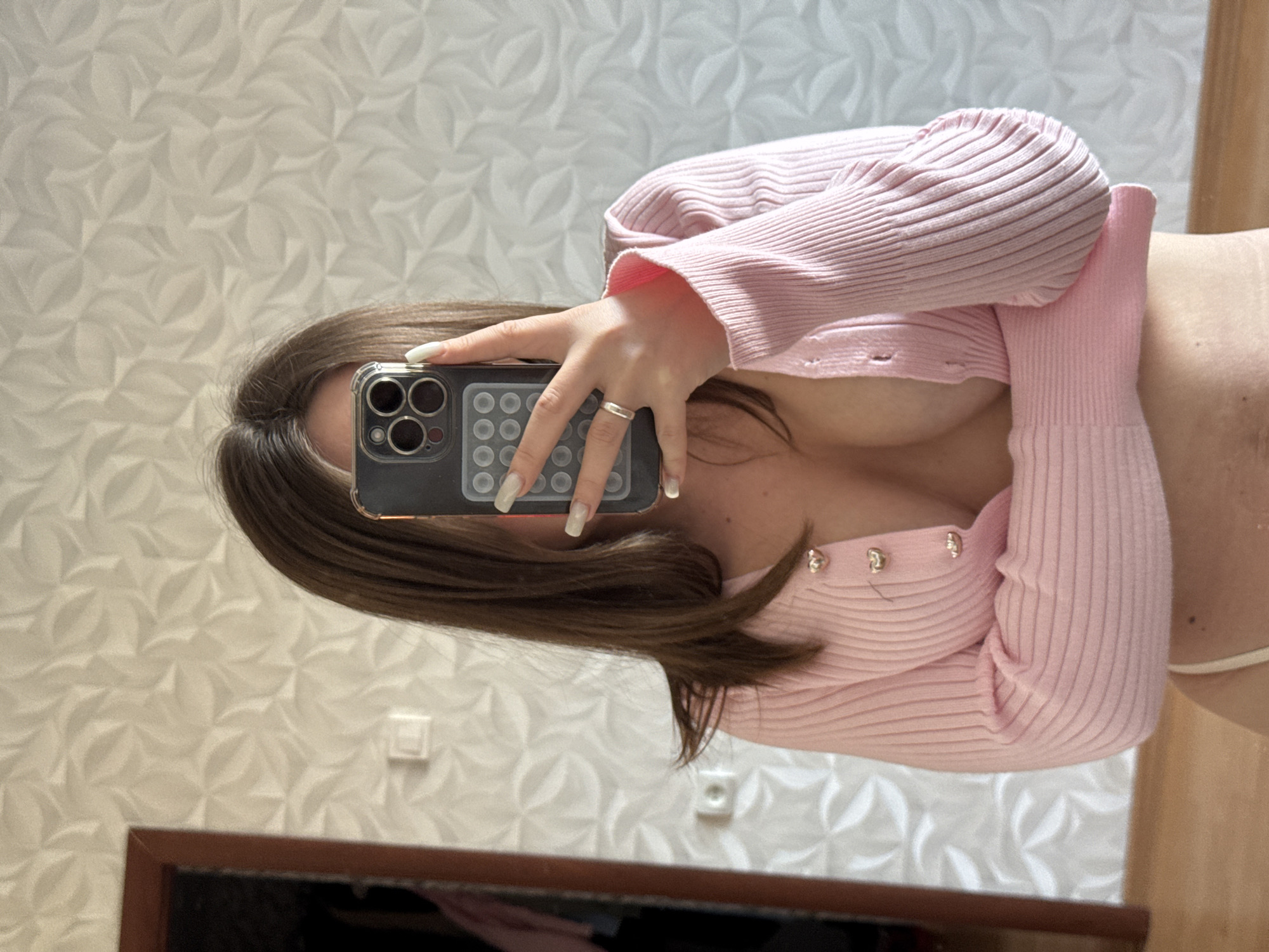 A woman with long brown hair is taking a mirror selfie. She is wearing a pink cardigan and has long, manicured nails. A silver ring is visible on her left hand. Her phone is black with multiple cameras. The background features patterned wallpaper and a partial view of a wooden-framed mirror. The image appears to be taken indoors. The location is stated as Orekhovo-Zuyevo, Russia.