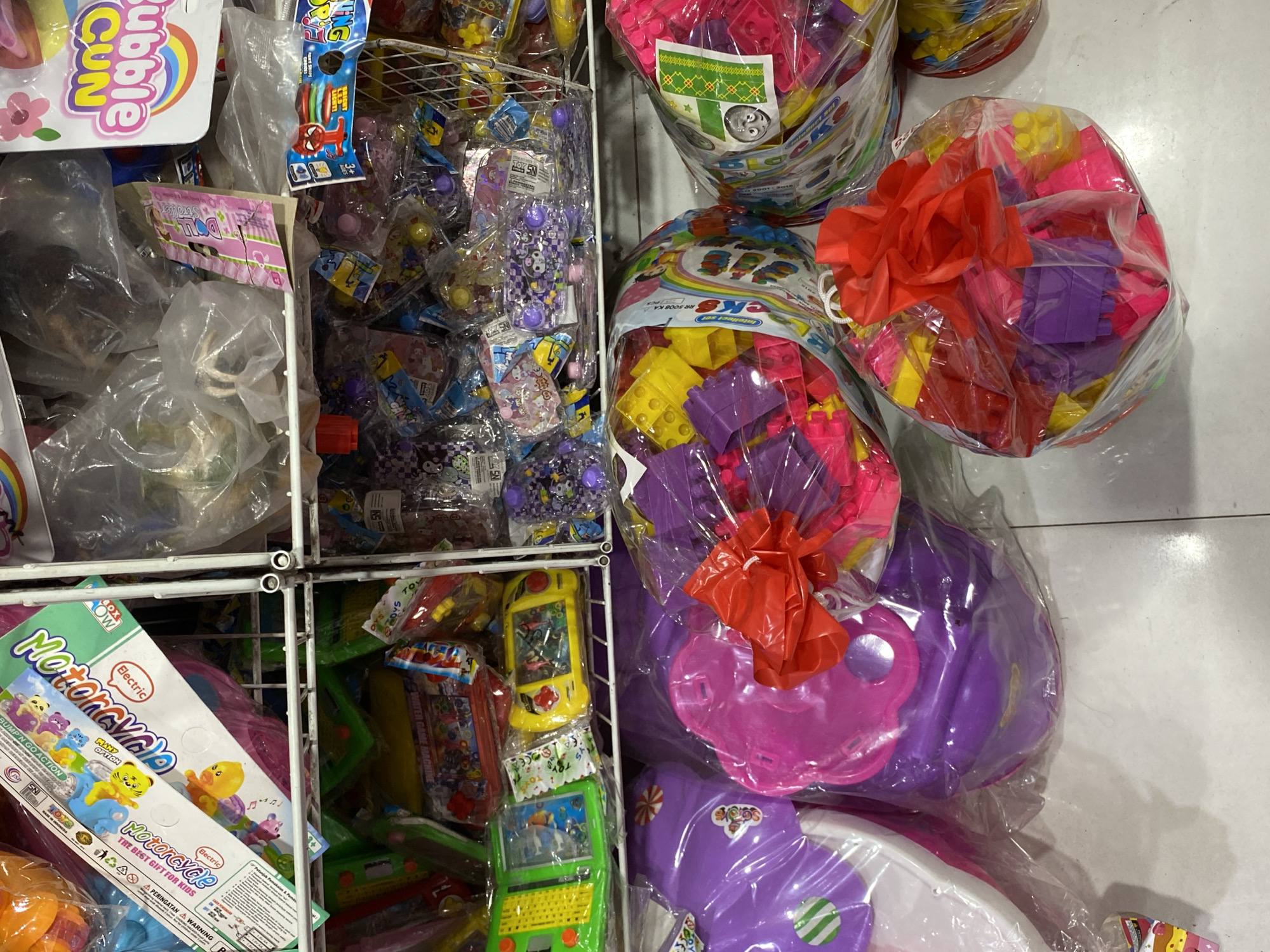 The image shows a retail display of toys, likely in a store or market stall in Banda Aceh, Indonesia. The toys are predominantly plastic and packaged in clear plastic bags, some with colorful branding.
In the foreground, several large, clear plastic bags contain colorful building blocks. One bag in the lower right corner is tied with a bright red ribbon. To the left, a large purple toy, possibly a play kitchen piece, is also packaged in clear plastic.
On the middle shelf, various smaller toys are visible. These include what appear to be water game machines in green and yellow cases, packaged toys with images of cartoon characters, and smaller plastic toys in brightly colored packaging. Some of these packages have text in Indonesian, such as "SNI" (Standar Nasional Indonesia) and "Mainan" (Toy).
The top shelf and beyond display more packaged toys, some in larger boxes with elaborate graphics, and a variety of plastic items in clear bags. The overall impression is of a busy, perhaps somewhat cluttered, toy retail environment. The lighting appears to be artificial, likely from overhead store lights, and the surfaces are clean and well-maintained. There are no people visible in the image.