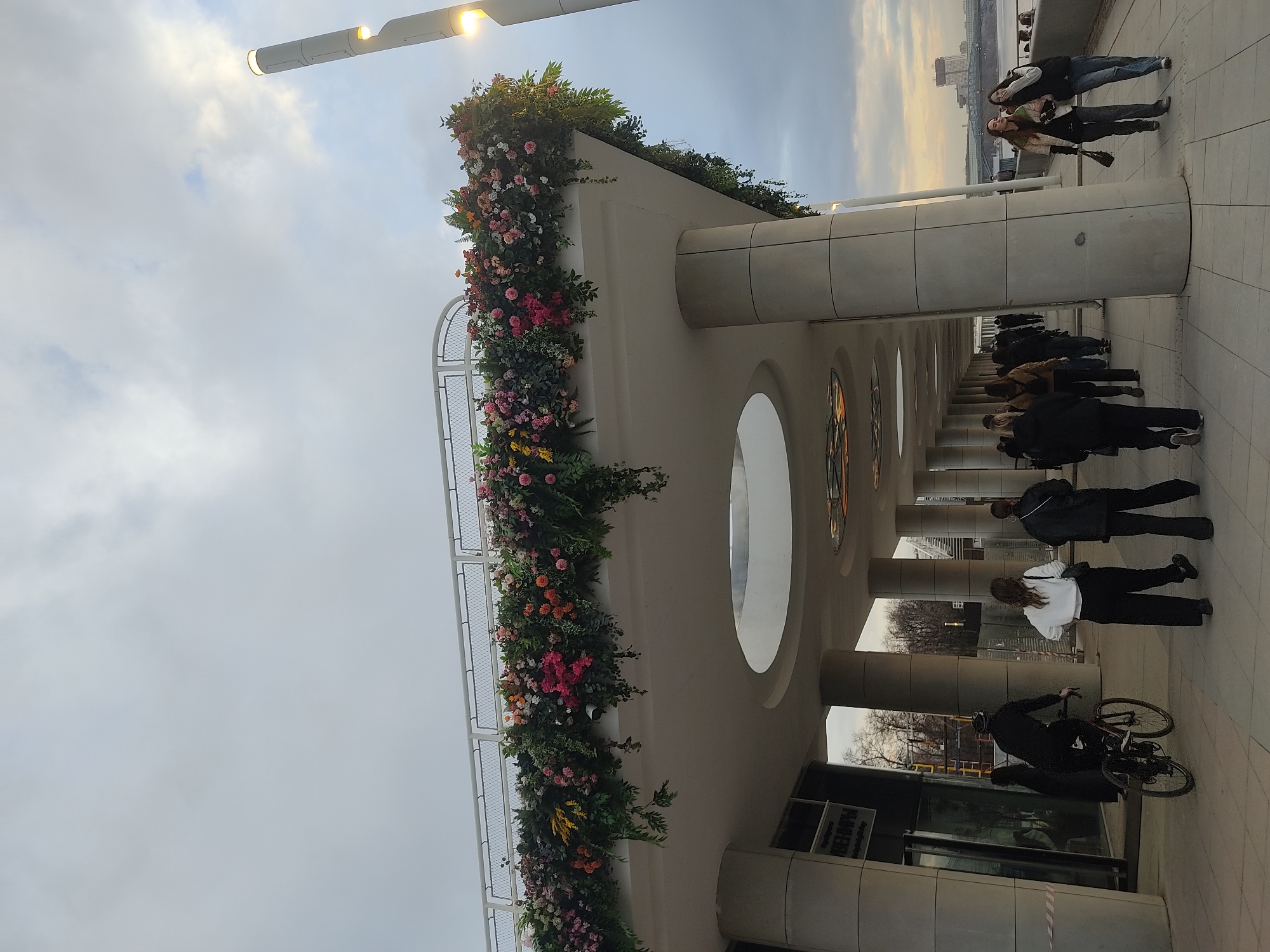 Here is a detailed description of the image:
The image shows an outdoor scene, likely in Moscow, Russia, given the architectural style reminiscent of the city's modern structures. The camera angle is tilted upwards, capturing a partially covered walkway adorned with vibrant floral arrangements on its upper section.
**Architecture and Environment**: The walkway is defined by a series of cylindrical columns, with large oval windows. The walkway is embellished with a cascade of flowers, spanning a variety of colors including reds, pinks, yellows, and greens. The sky above is overcast, with a gradient of light fading into the horizon.
**People and Activity**: The photo captures a diverse group of people. Several pedestrians are walking along the walkway, some appearing to be in casual attire. Notably, one individual is riding a bicycle through the area.
**Additional Details**:
-In the background, the distant cityscape and a river are visible, indicating a location near a body of water.
-A modern lamppost with an unusual shape is positioned near the top of the image.
-A store or business is on the bottom of the frame.
-The environment looks like the transition to Autumn.
Overall, the image depicts a lively scene within a modern architectural setting in Moscow.