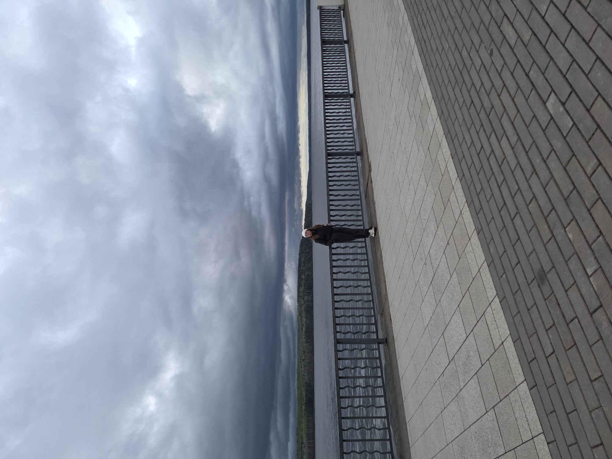 A person stands on a pedestrian walkway along a riverfront in Chaikovsky, Russia. The person is facing toward the camera with their back to a metal railing which runs parallel to the river. The walkway is paved with light gray pavers, and on the right is a dark gray brick walkway. The river appears to stretch to the horizon. The sky is filled with dark, ominous clouds.