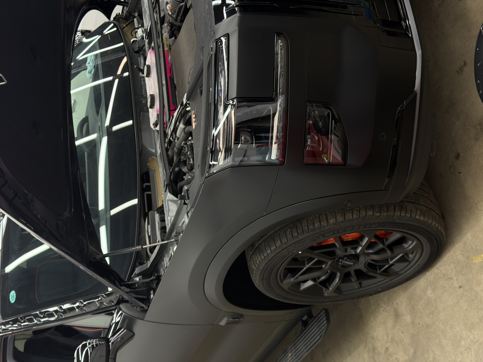 A matte black SUV with its hood open is parked on a concrete floor, likely inside a garage or workshop. The vehicle features black rims with orange brake calipers. The engine compartment is visible with various components. The car is angled with its front end facing the right side of the image. The location of the image is not discernible from the visual information provided. The city of Krasnoyarsk, Russia is not evident in the image. There are no people visible.