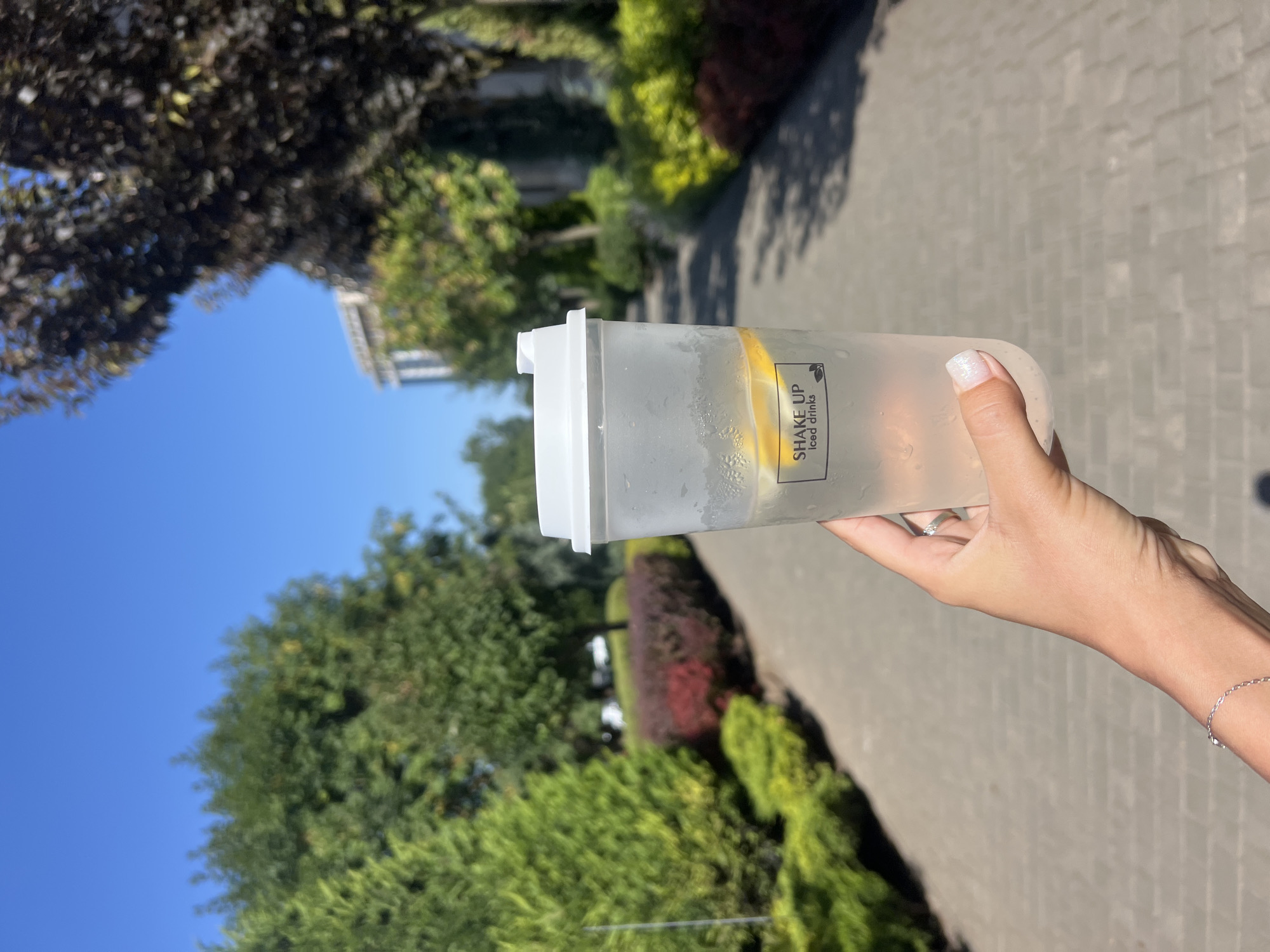 A woman's hand holds a frosted plastic cup with a white lid filled with a clear beverage and lemon slices. The cup has the text "SHAKE UP iced drinks" printed on it. She is wearing a silver ring and a delicate silver bracelet. The background is a paved walkway lined with manicured green bushes and trees, with a building visible in the distance. The sky is clear blue. The scene appears to be outdoors, likely in a park or well-maintained public area within the city of Fontanka, Ukraine.