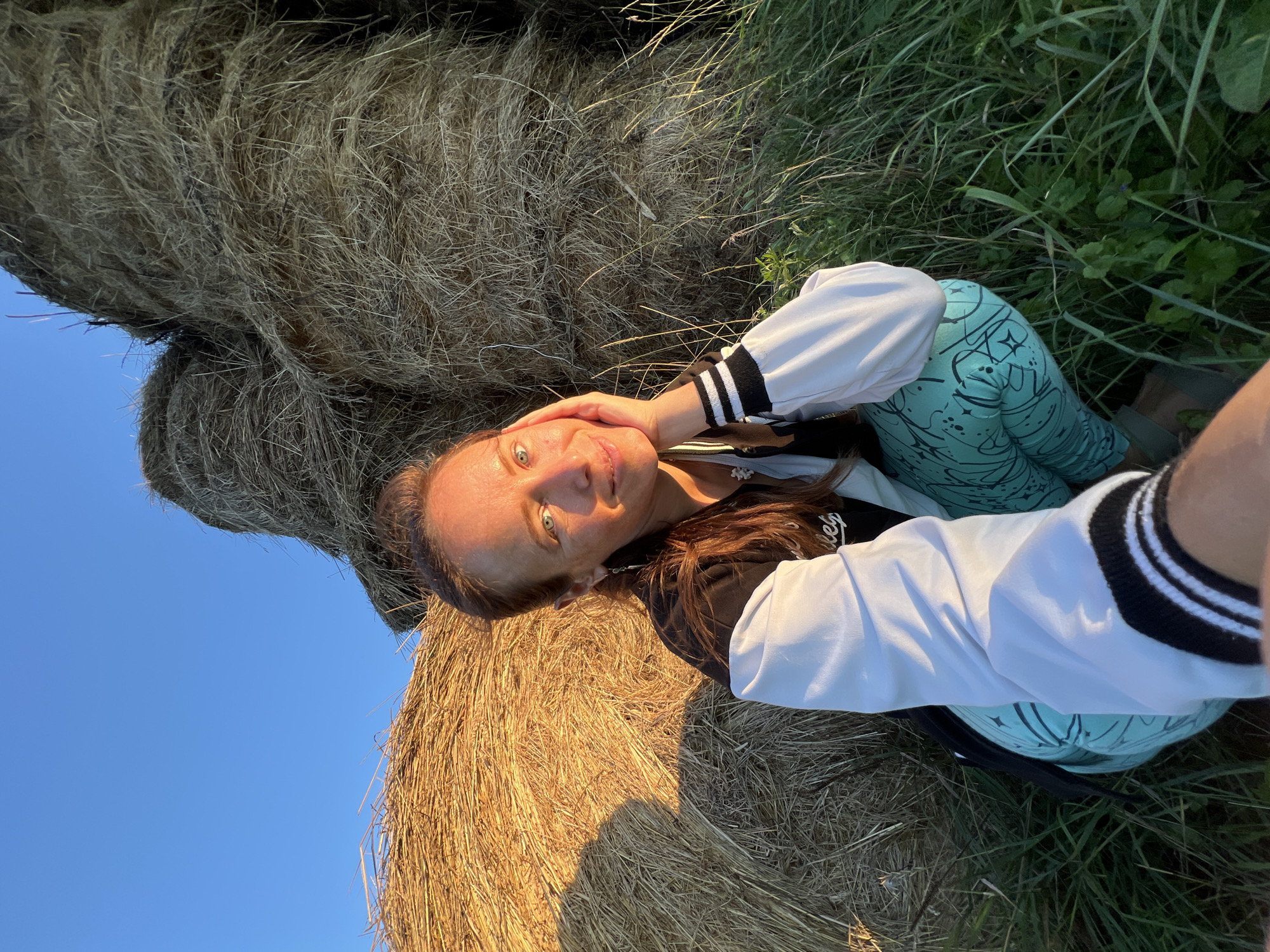In Pivkino, Russia, a woman with auburn hair, wearing a black and white jacket and teal patterned leggings, is captured in a selfie. She is crouching in front of large hay bales, bathed in the warm glow of what appears to be late afternoon sun. The sky above is a clear blue. She rests her right hand on her cheek, looking directly at the camera with a gentle smile. The foreground is filled with green grass and foliage, suggesting an outdoor, rural setting.