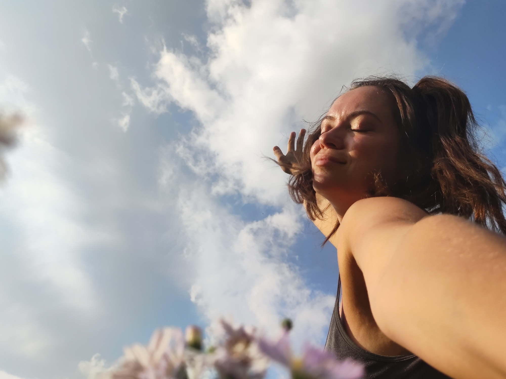 A low-angle, wide shot shows a young woman with her eyes closed, head tilted back and a serene expression, bathed in sunlight. She is wearing a dark-colored tank top. Her right arm is extended towards the camera, with her hand blurred and obscuring part of the frame. Her hair is pulled back into a ponytail. In the foreground, out-of-focus pink and purple flowers are visible. The background is a bright blue sky with scattered white clouds, suggesting an outdoor setting. The image does not provide any specific details about the city of Фоки, Russia.