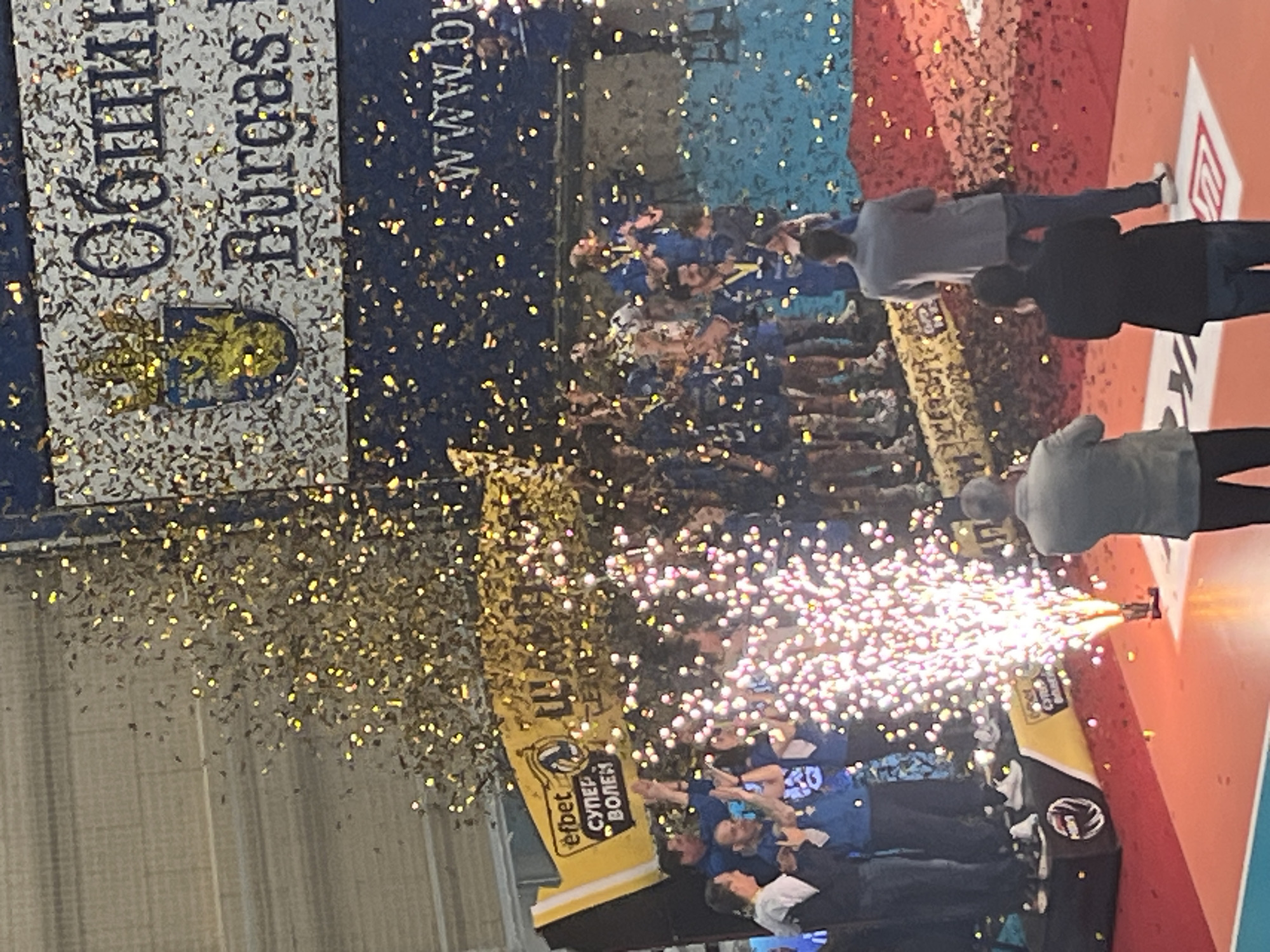 The image depicts a celebration, likely a sports victory, taking place in what appears to be an indoor arena in Burgas, Bulgaria. Confetti is being released into the air, and pyrotechnics are being set off. A large group of people, presumably the winning team and associated personnel, are gathered on a platform. The banner for "Neftochimik 2010 Burgas" is prominent. Spectators are also visible. The background shows branding and advertising elements. Some individuals are observing and likely recording the event.