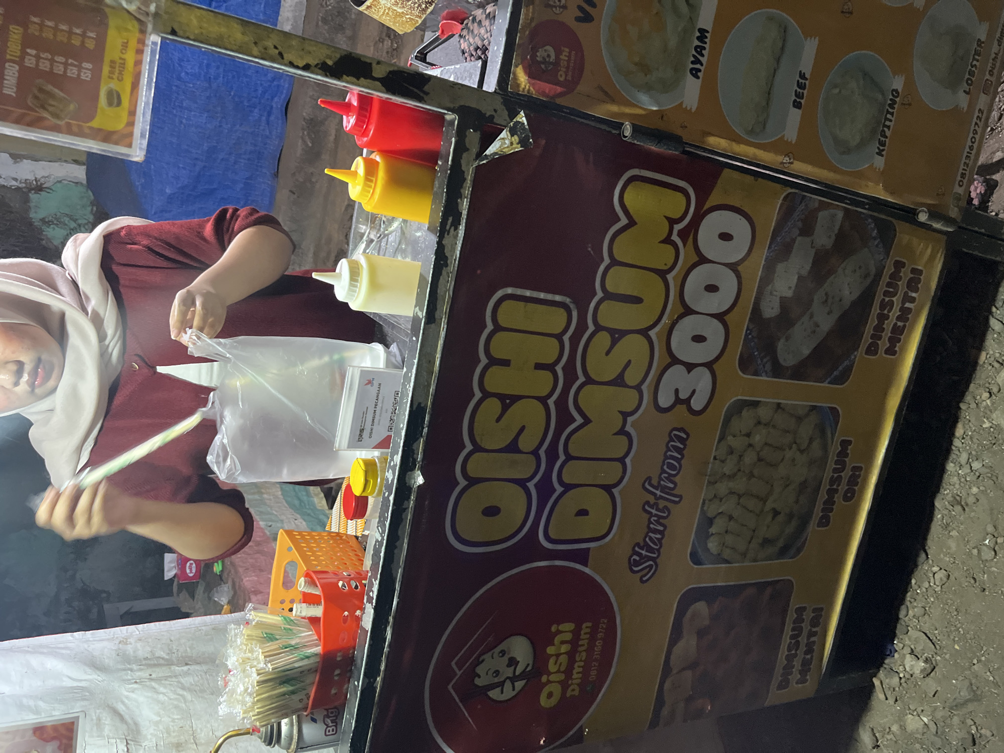 The image shows a street food stall in Pecangaan, Indonesia, selling dim sum. The stall is decorated with a large banner that says "OISHI DIMSUM" and "Start from 3000". There are pictures of different dim sum varieties on the banner, including mentai, original, chicken, beef, crab, and lobster. A person, wearing a headscarf and a maroon top, is seen preparing food behind the counter. Various condiments, including ketchup, chili sauce, and mayonnaise, are also visible. The stall appears to be set up on a street with uneven ground, suggesting it is an outdoor market or roadside vendor. The lighting suggests it is evening or night.