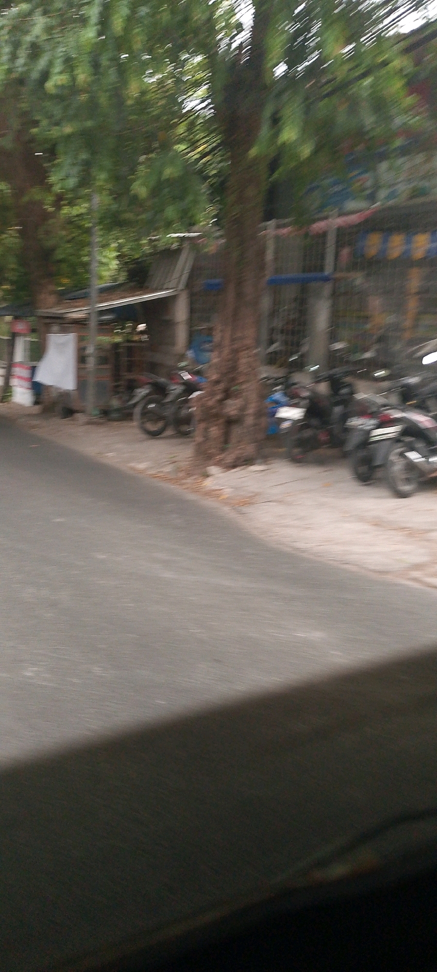 A slightly blurred, eye-level, outdoor, medium shot shows a row of parked motorcycles on a sidewalk in front of a building. The building appears to be a small shop or stall with a corrugated metal roof and a white cloth hanging from its front. The motorcycles are parked along the sidewalk, some with their headlights on. A large tree with a thick trunk stands in the foreground, partially obscuring the building and motorcycles. The sidewalk is made of concrete. A paved road runs along the left side of the frame. The background is filled with green foliage from trees. The scene is likely a street in a residential or commercial area of Bandung City, Indonesia, judging by the presence of motorcycles, the style of the building, and the tropical vegetation. The lighting suggests it is daytime.