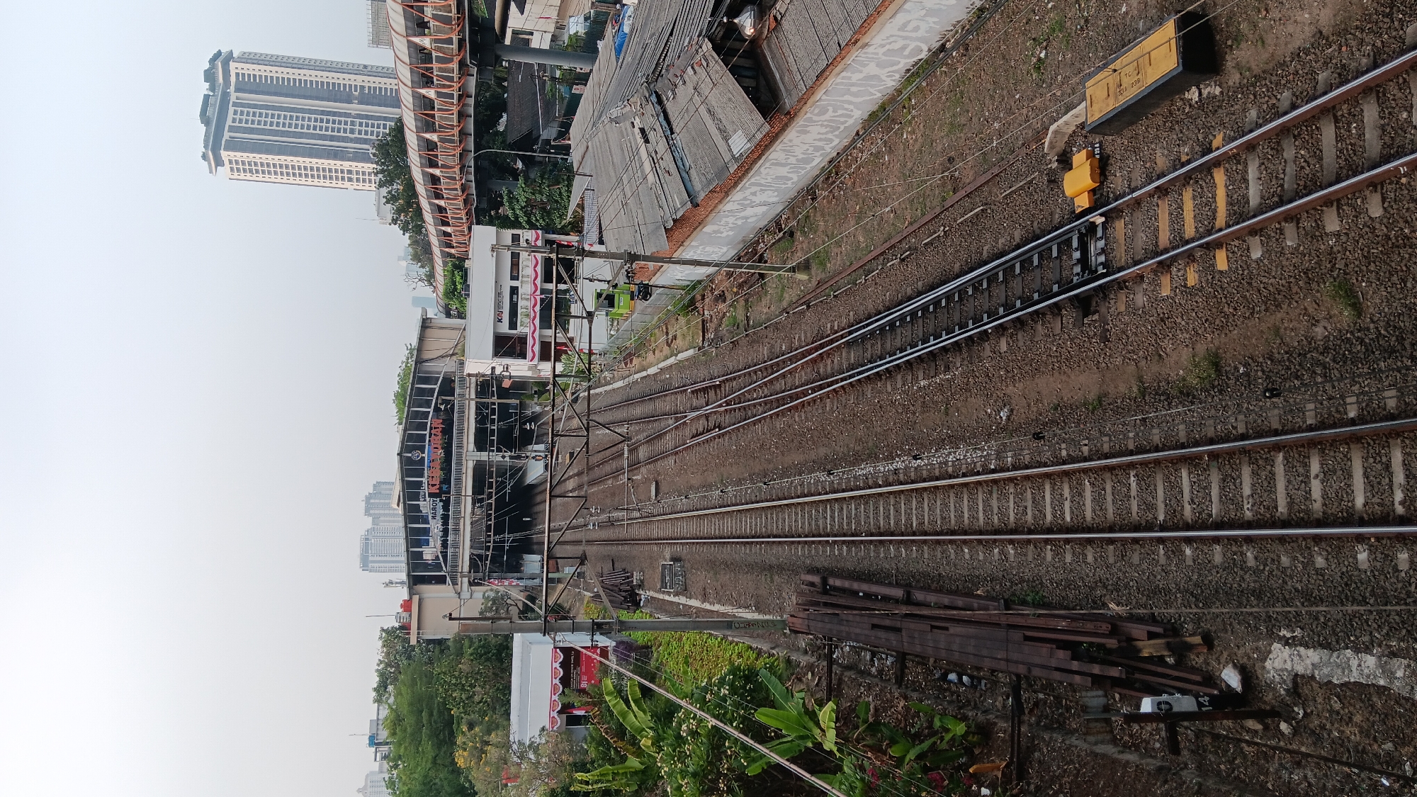 The image depicts a train station and railway tracks in South Jakarta, Indonesia. In the foreground, multiple sets of train tracks converge and extend into the distance. To the left of the tracks, a pile of wooden railway ties is stacked. A white pole with a black marker labeled "14" is visible near the ties.
Further back, a train station building is visible with a prominent sign reading "MAJOT KESEDRERAN". The station appears to be elevated, with an overpass or bridge spanning across the tracks in the background. The bridge has an orange railing and is enclosed. A tall, modern building with many windows stands prominently in the background, characteristic of urban development in Jakarta.
The environment is somewhat hazy, suggesting the urban atmosphere or possibly a cloudy day. Vegetation, including trees and plants, is present on the left side of the image. The rooftops of several other buildings, some with corrugated metal, are visible to the right of the tracks, indicating a densely populated urban area. There are no people visible in the image. The scene is primarily a landscape of urban infrastructure related to the railway system.