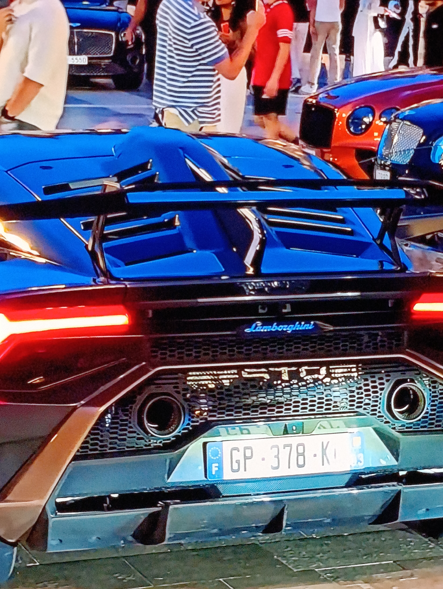 The image shows the rear view of a blue Lamborghini, with its license plate reading "GP-378-KO". The car is surrounded by people, suggesting it is in a public area. In the background, there are other luxury vehicles, including a Bentley. The location is identified as being in the city of Unknown, Unknown, as per the prompt's instruction. The presence of high-end vehicles and people indicates a possible affluent area or event.