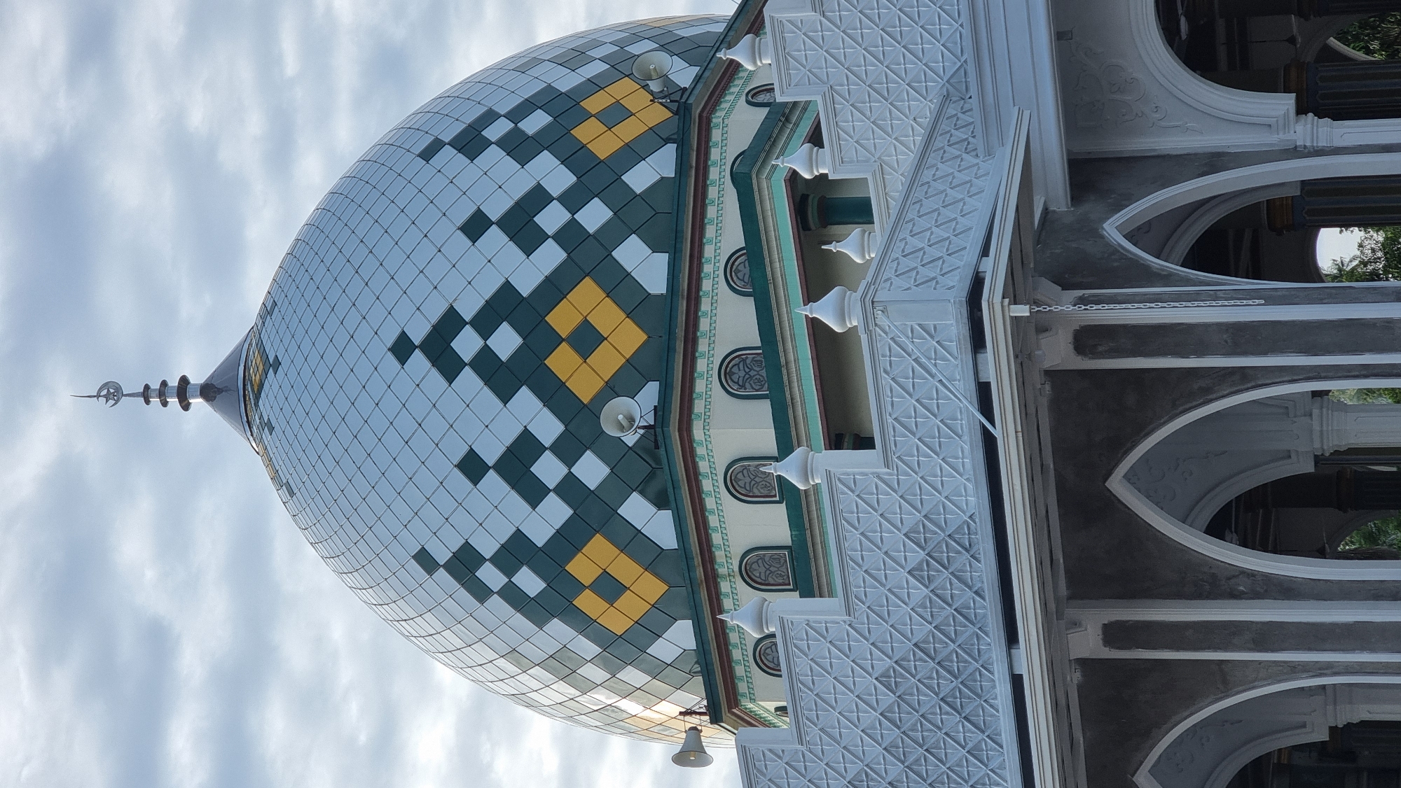 The image shows a close-up, eye-level view of the dome and upper facade of a mosque in Meunasahlhok, Indonesia. The dome is large and multifaceted, covered in a pattern of diamond-shaped tiles that alternate between light silver, dark green, and yellow. A crescent moon and star finial tops the dome. Below the dome, the mosque has a pale yellow facade with arched windows and decorative green trim. In the foreground, white decorative railings and arches of a lower structure are visible. Two white loudspeakers are mounted on the sides of the dome. The sky above is overcast with light grey clouds. There are no people visible in the image.