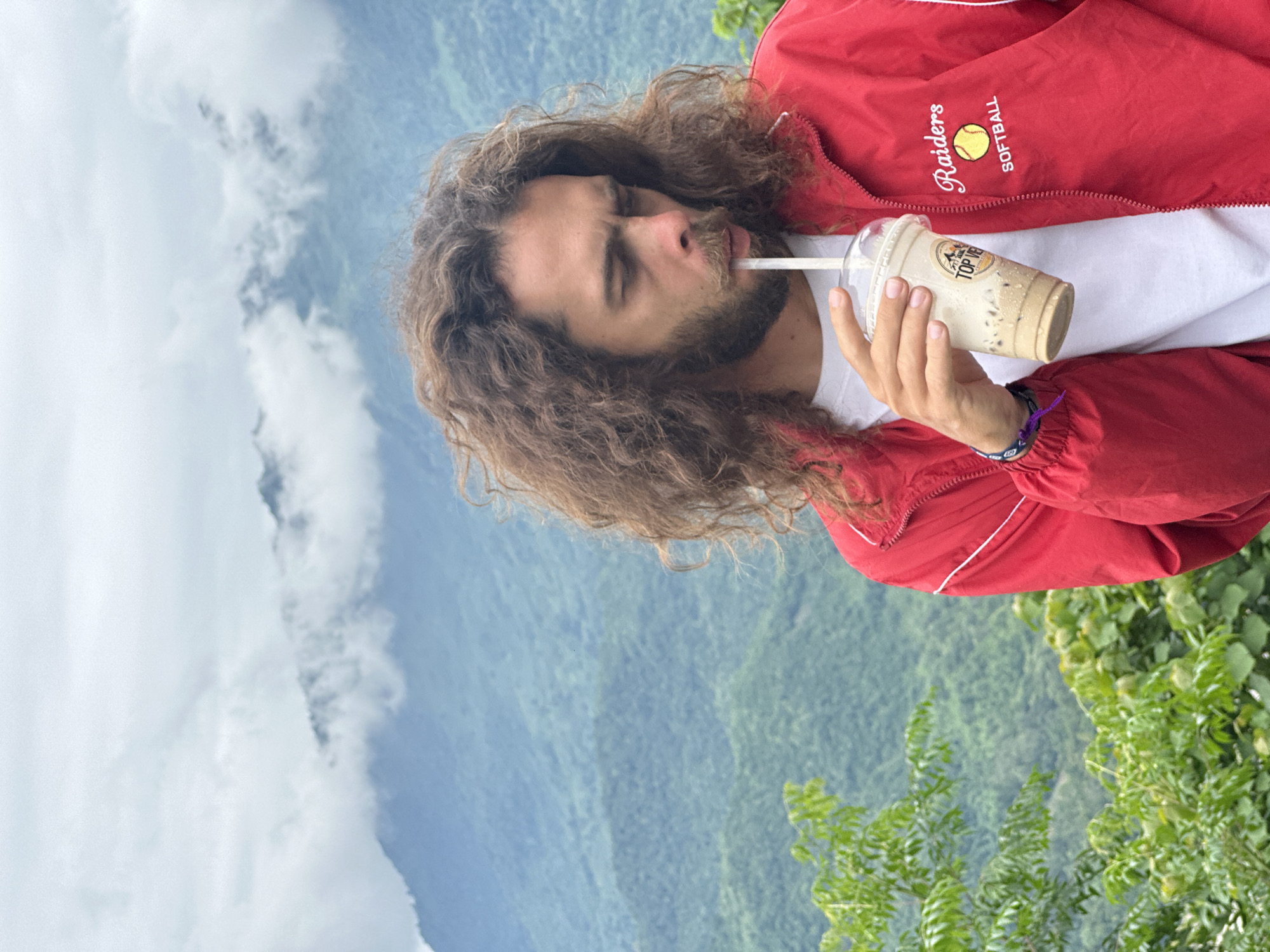 A person with long, curly brown hair and a beard is drinking from a plastic cup of iced coffee. They are wearing a red jacket with "Raiders SOFTBALL" and a yellow softball logo embroidered on the left chest. The person is standing outdoors, with a backdrop of lush green mountains and clouds. The scene appears to be from a viewpoint in Mai Chau, Vietnam, given the characteristic mountainous terrain and vegetation. The person is holding the cup with their right hand, and a straw extends from the cup to their mouth. A blue and purple beaded bracelet is visible on their left wrist. The lighting suggests it is daytime, with an overcast sky.