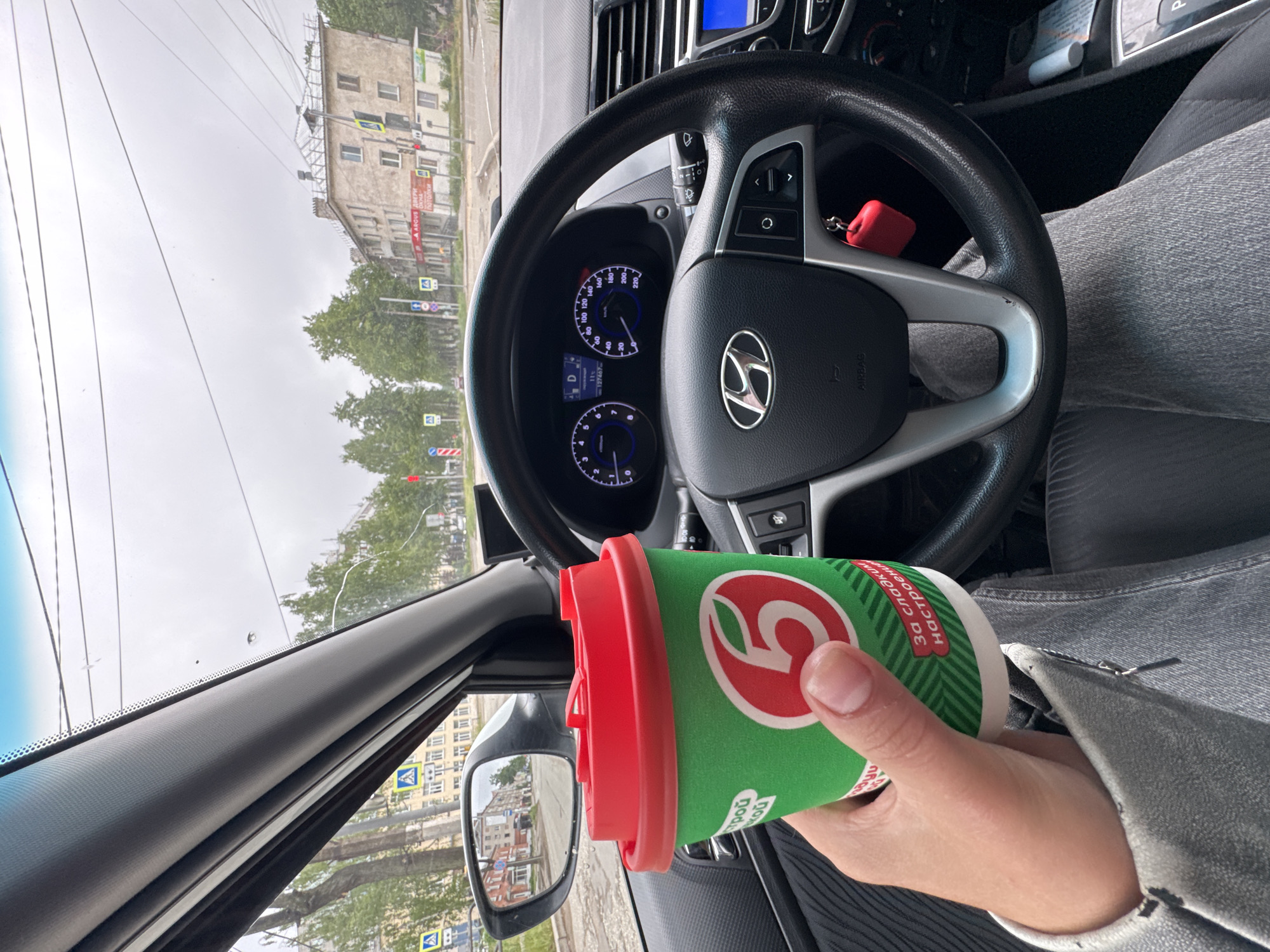 The photo, taken from the driver's seat of a Hyundai, shows a person holding a green and white coffee cup with a red lid bearing the "5" Pyaterochka logo. The steering wheel and dashboard of the car are visible, and outside the car window is a street scene with buildings and trees.
Based on the buildings and traffic signs visible through the car windows, this photo was taken in the city of Chaikovsky, Russia.
