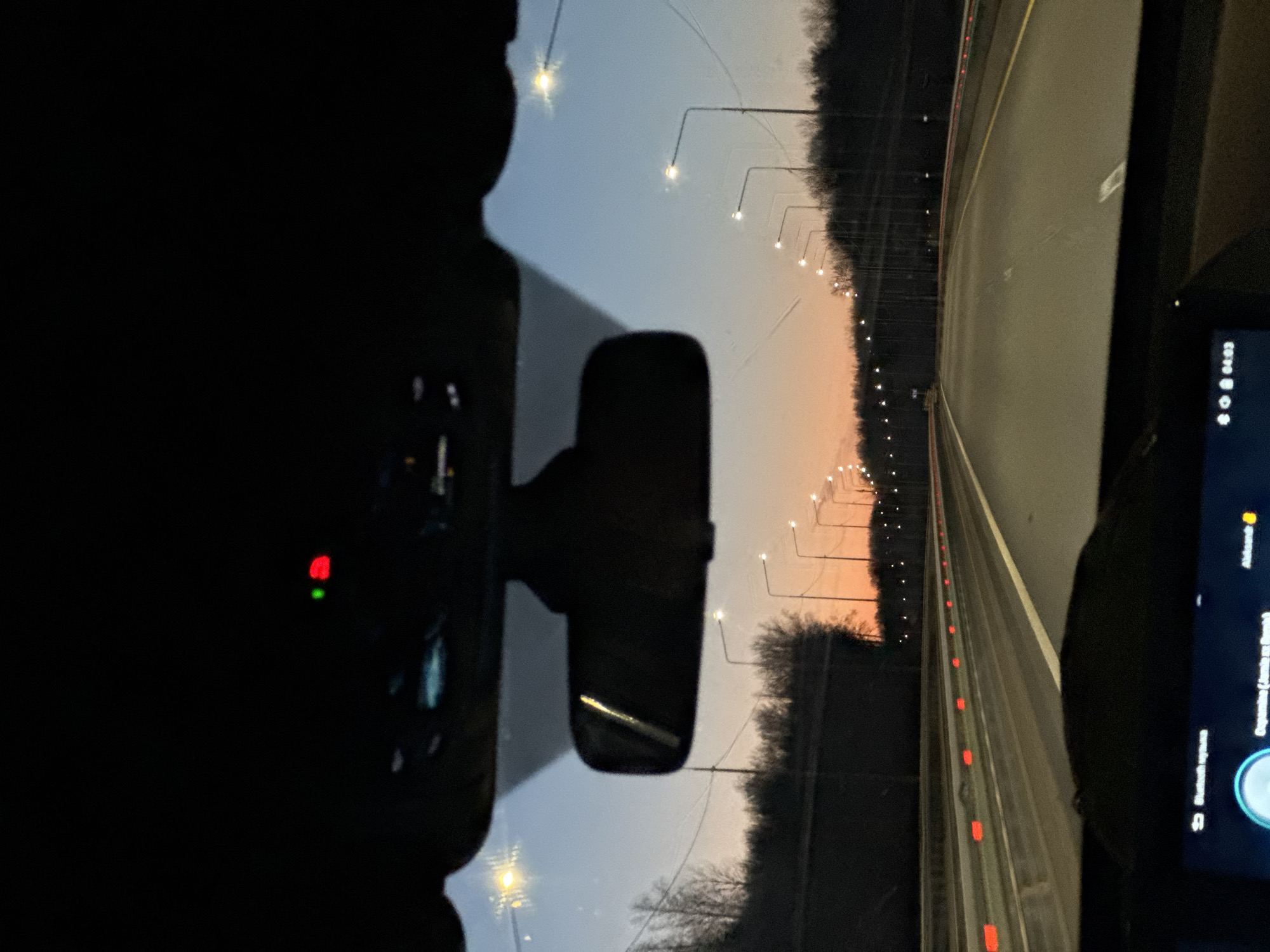 The image shows a view from inside a car driving on a highway at dusk or dawn. Streetlights line both sides of the road, receding into the distance. The rearview mirror is visible, and a dashboard screen displays information. The sky is a gradient of blue and orange. The location is unknown.