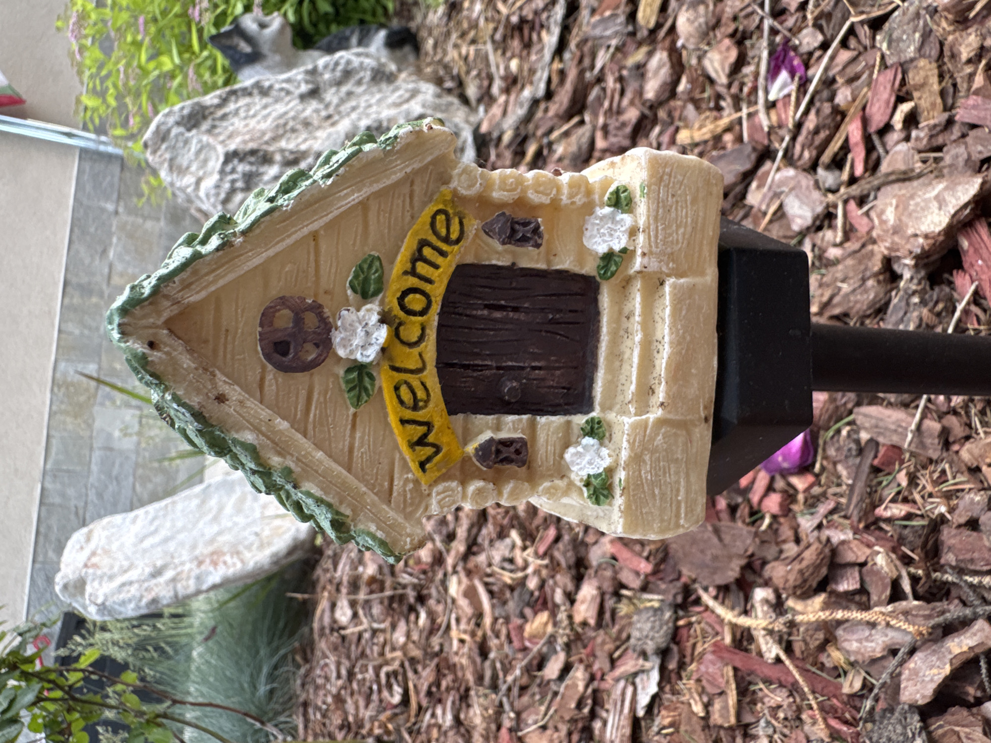 A top-down shot shows a miniature "Welcome" birdhouse attached to a black metal post, set on a bed of reddish-brown mulch. The birdhouse is tan with a brown door and a yellow "Welcome" sign. The yard is located in the city of Unknown, Unknown.