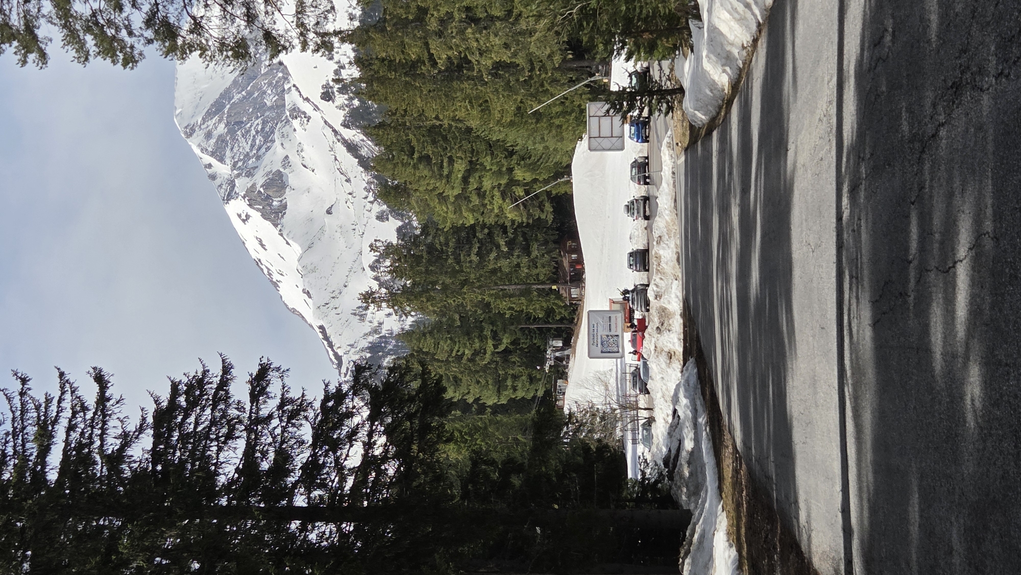 The media file is a photograph of a snowy mountain landscape, presented rotated 90 degrees clockwise.
**Subjects:** The central subject is a large, snow-covered mountain with rocky outcrops, dominating the left side of the frame when viewed correctly. A dense forest of dark green coniferous trees (pines) stretches across the midground. In front of the trees, several cars and SUVs, mostly dark-colored, are parked on a snow-covered area. The foreground features a paved asphalt road lined with patches of snow and bare earth. No people are visible.
**Activity or event taking place:** The scene suggests a winter or early spring day at a mountain resort or scenic area, likely associated with skiing or other cold-weather activities given the snow and mountain backdrop. The parked cars indicate visitor presence.
**Notable details:** The time of day appears to be mid-morning to early afternoon, evidenced by the long, distinct shadows cast by trees across the road. The sky is a pale blue with some light cloud cover. The environment is mountainous and snowy.
**Visible text:** On a white sign located near the parked cars, a QR code is prominently displayed. Accompanying text includes "frystok.cz", "ski-areal.cz", and "Instagram". The word "FRENŠTÁT" is also faintly visible on a smaller sign element.