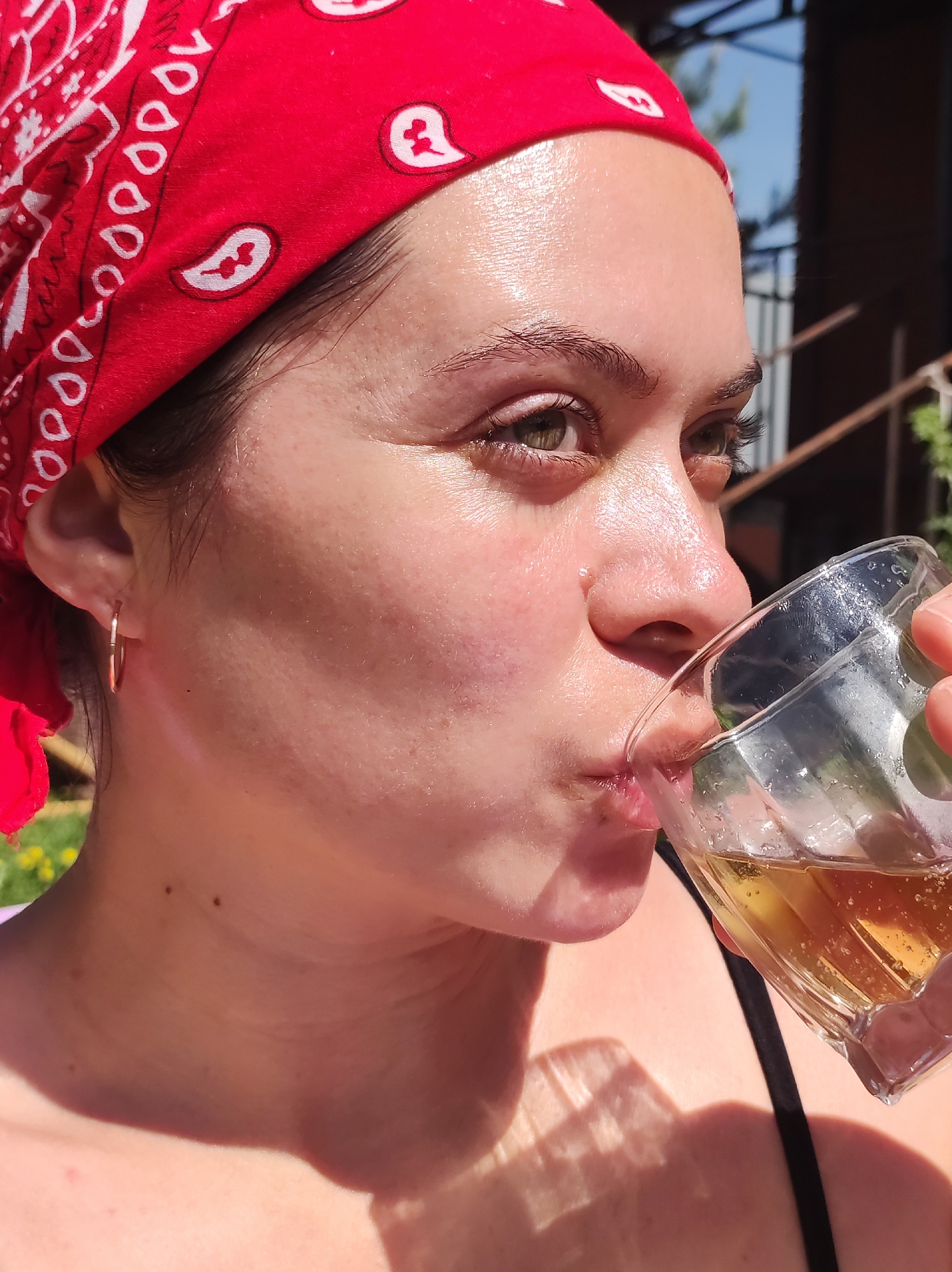 A woman with fair skin and wearing a red bandana with white paisley designs is drinking from a glass of amber-colored liquid. She has green eyes, dark eyebrows, and a gold hoop earring. The woman's face is partially lit, suggesting strong sunlight. The location is possibly in Фоки, Russia, but it is impossible to confirm without further information. The environment appears to be outdoors with possible structures or vegetation in the background. The woman is taking a drink, the glass is close to her mouth.