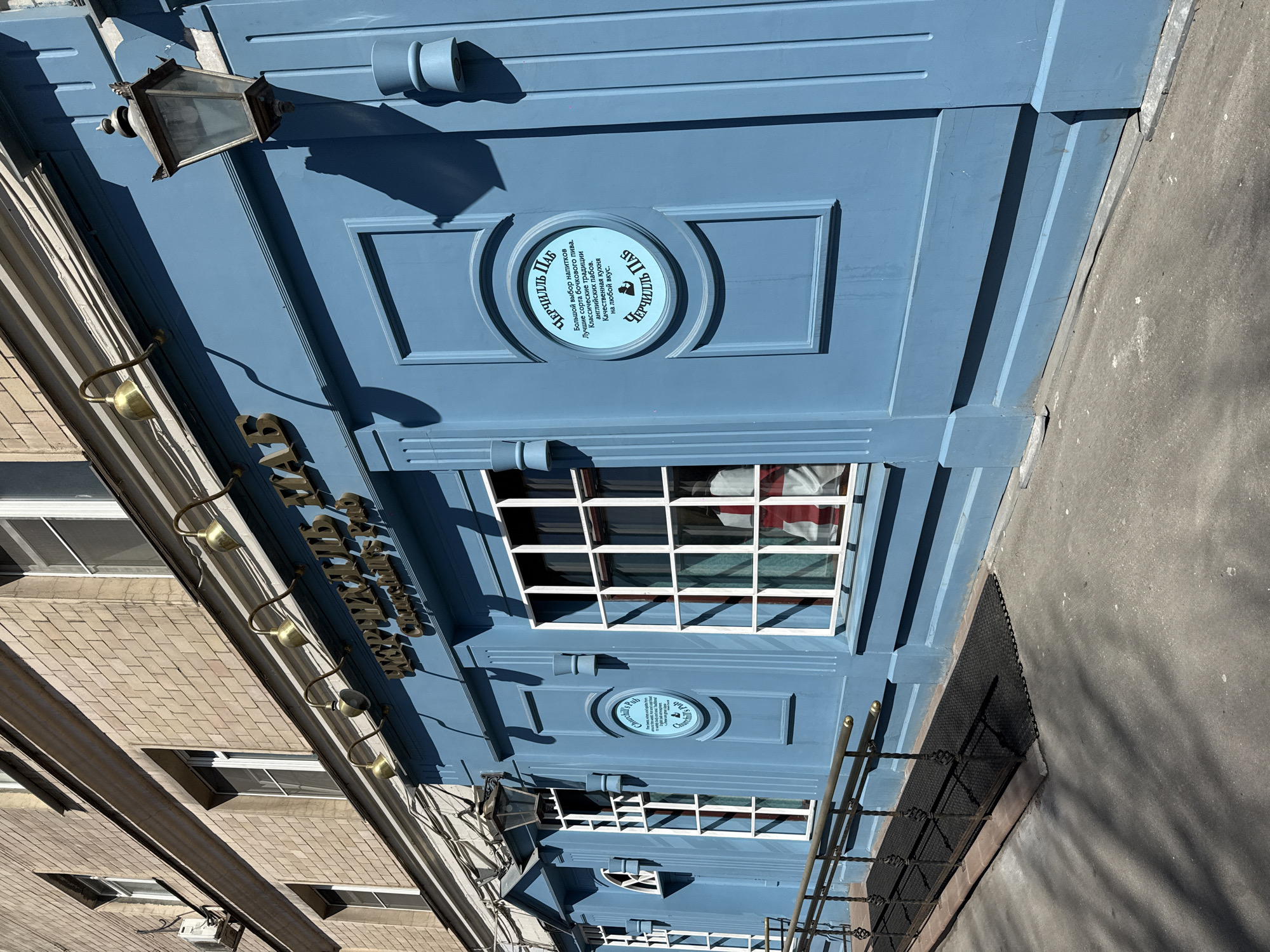 Here is a forensic description of the image:
The image shows the exterior of a building in Khimki, Russia, identifiable by signage and architectural style. The building's facade is painted blue and features detailed molding. Signs reading "Churchill Pub" are visible in both Russian and English. A window with a white grid frame has a partial view of what appears to be a Union Jack flag inside. An old-fashioned lantern is mounted above the windows, and several decorative brass fixtures are spaced along the side of the building. A brick building stands next to the pub. The setting appears to be a street or urban area.