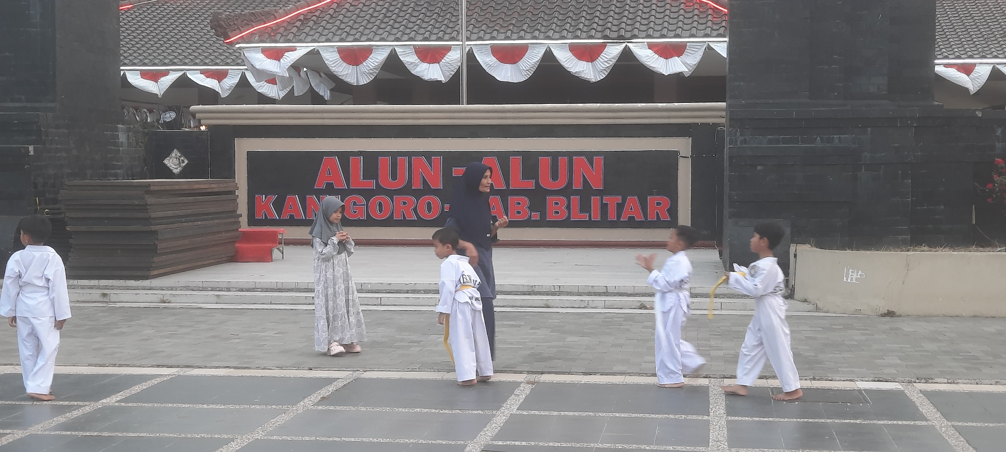 In the city of Kanigoro, Indonesia, a group of children dressed in white martial arts uniforms are gathered outside a building with a sign that reads "ALUN-ALUN KANGORO-AB. BLITAR". A woman in a dark headscarf, likely an instructor, stands among the children. The children appear to be participating in a training or demonstration session, with some in motion as if practicing their moves. The environment is an outdoor public space with paved ground and decorative banners on the building.