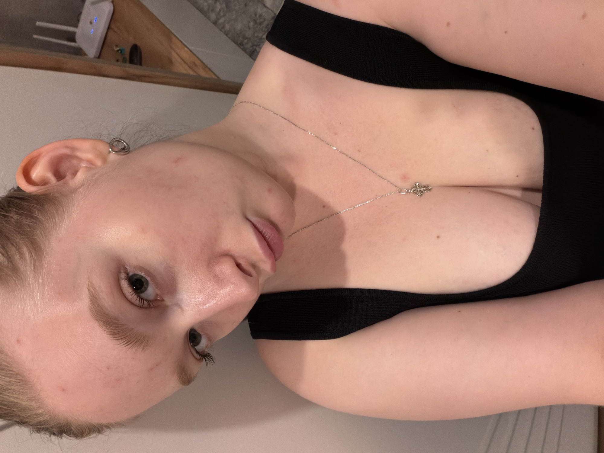 A young woman with blonde hair pulled back is leaning against a white wall, looking down and to her right. She is wearing a black tank top with a silver cross pendant. Her left ear has a silver hoop earring. The background is blurred, showing a wooden shelf with a white router. The setting appears to be indoors, possibly a room in a home. There is no indication of Rossiyskiy, Russia in the image.