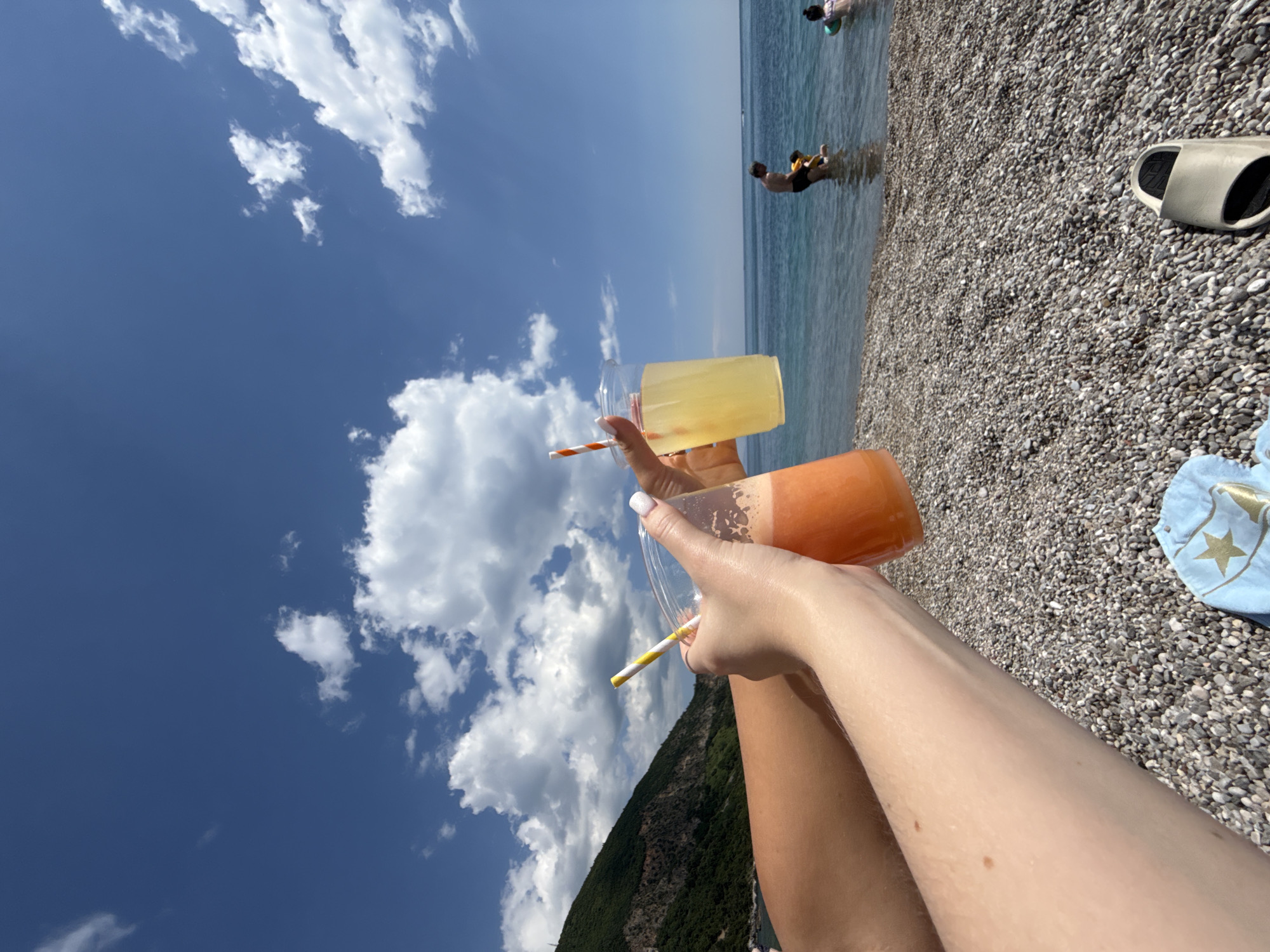 A first-person view shows a person holding two drinks on a pebble beach in Prijevor, Montenegro on a sunny day. The drinks are in clear plastic cups with striped straws; one is yellow, the other orange. The person's hand and legs are visible. In the background is the Adriatic Sea, with two people wading in the water. The sky is blue with scattered white clouds. To the right are gray-and-black slides and a light blue towel with a gold star pattern on the pebble beach. A green, forested hill slopes down towards the sea on the lower left.