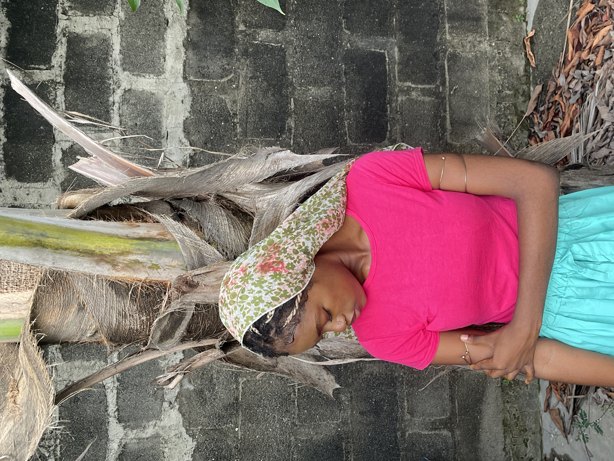 A young person is depicted lying horizontally on their side, seemingly resting or sleeping, against the base of a large, fibrous plant trunk, likely a palm tree. The individual, who appears to be female and has dark skin, is wearing a vibrant pink t-shirt and a light blue or turquoise skirt. A patterned headscarf, featuring green, white, and pink floral designs, covers their head. Their eyes are closed, and they have a small nose stud and multiple gold-colored bangles or armbands on their upper arms. Their hands are clasped or intertwined over their stomach area.
The setting is outdoors, with a rough, weathered concrete block wall forming the background. The ground or surface around the plant base is covered with dried leaves, debris, and other natural textures from the plant itself. The lighting suggests it is daytime, although specific time of day or weather conditions cannot be definitively determined. The overall atmosphere conveys a moment of quiet repose. No visible text is present in the image. The environment is consistent with an outdoor, potentially semi-rural or residential area in Ado, Nigeria.