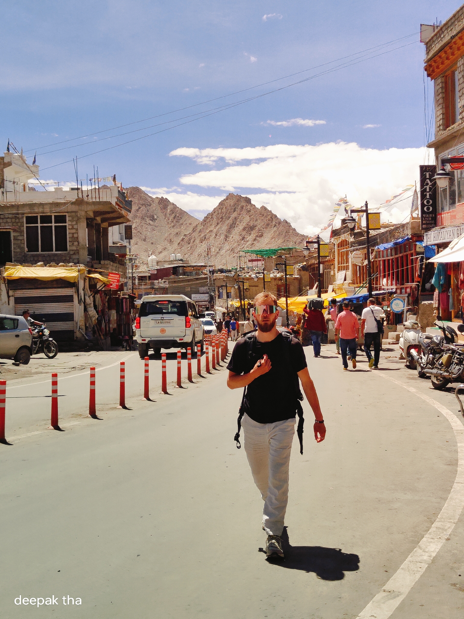 The image depicts a street scene in Leh, India, likely during the daytime, with a bright, sunny sky and some clouds. The main subject, a man wearing sunglasses, a black t-shirt, and light-colored pants, is walking towards the camera on the right side of the road. He is carrying a backpack. The street is lined with buildings on both sides, featuring shops and stalls. Various vehicles, including a white SUV and a motorcycle, are visible on the road. People are seen walking along the street and browsing in the shops. Mountains form the backdrop to the urban environment. Prayer flags are strung across some of the buildings, indicating a Tibetan Buddhist cultural influence. The overall atmosphere appears to be that of a bustling market or commercial area in a mountainous region.