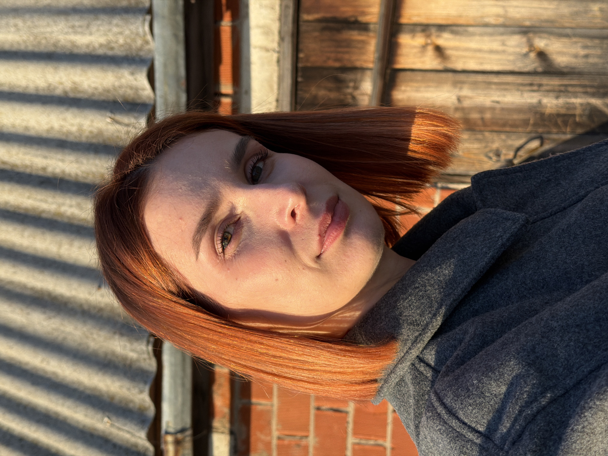 A close-up portrait of a fair-skinned woman with shoulder-length auburn hair. She is wearing a gray wool coat. The background is a combination of corrugated metal, weathered wooden planks, and red brickwork. The lighting suggests an outdoor setting. The location is Chaikovsky, Russia.