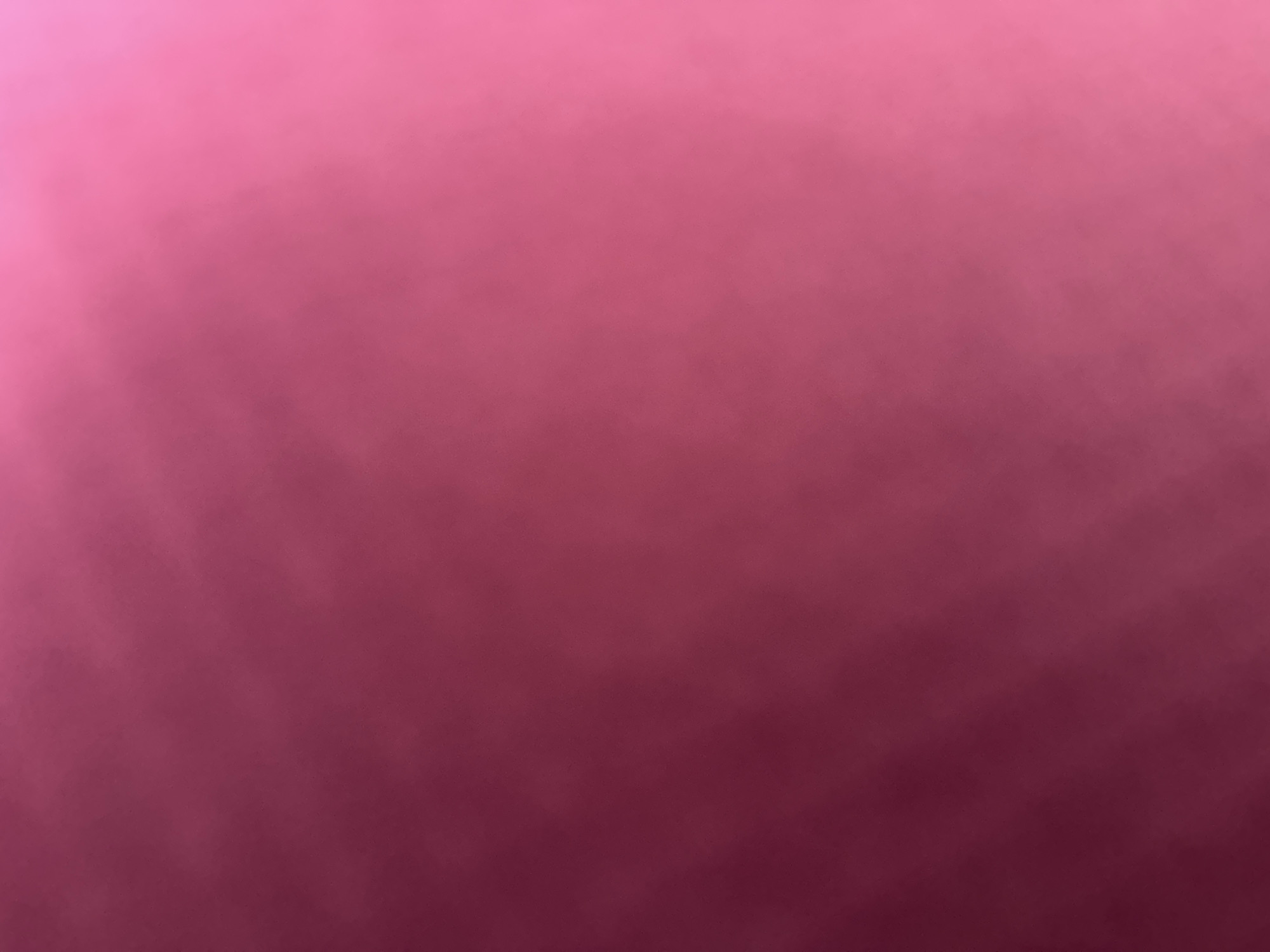 The image is a photograph with a soft, blurry background in shades of pink and magenta. There are no discernible subjects, objects, or activities taking place. The lighting appears to be diffused, creating a gradient of color across the frame, with the top left corner being lighter and transitioning to a deeper magenta at the bottom right. There are no visible signs of a specific time of day, weather conditions, or location cues. The overall impression is abstract due to the lack of focus and clear detail. There is no visible text in the image.