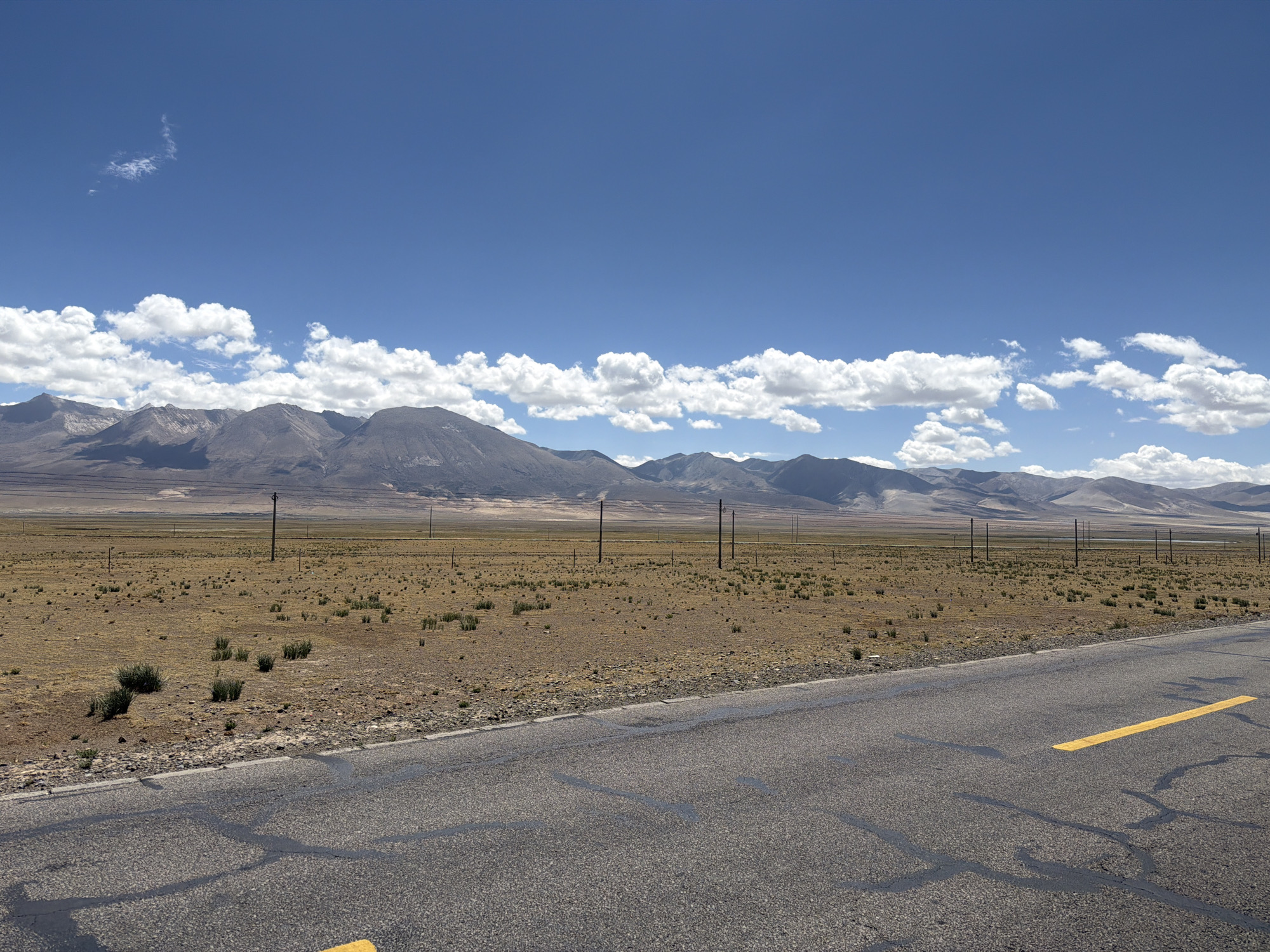 The image displays a vast and open landscape under a clear blue sky, characteristic of Mainpu township, China. In the foreground, a two-lane paved road runs diagonally from the lower left to the middle right, featuring visible cracks in its asphalt surface and a bright yellow dividing line along its edge.
To the left of the road, a wide, flat plain extends towards distant mountains. This terrain is predominantly dry and light brown, sparsely covered with small, low-lying green and brownish bushes. A series of dark utility poles, possibly for power or communication, are seen stretching across the plain into the distance, adding a man-made element to the natural expanse.
In the background, a long range of rugged, treeless mountains rises, their peaks appearing grey and brown under the bright sunlight. Above the mountains, the sky is a vibrant blue, adorned with scattered, fluffy white cumulus clouds. A lone, wispy cloud is visible higher in the sky to the left.
The scene is bathed in bright daylight, indicating clear weather. The overall impression is one of immense space, solitude, and the raw beauty of a high-altitude or arid environment. There are no people, animals, or visible text other than the road marking.
