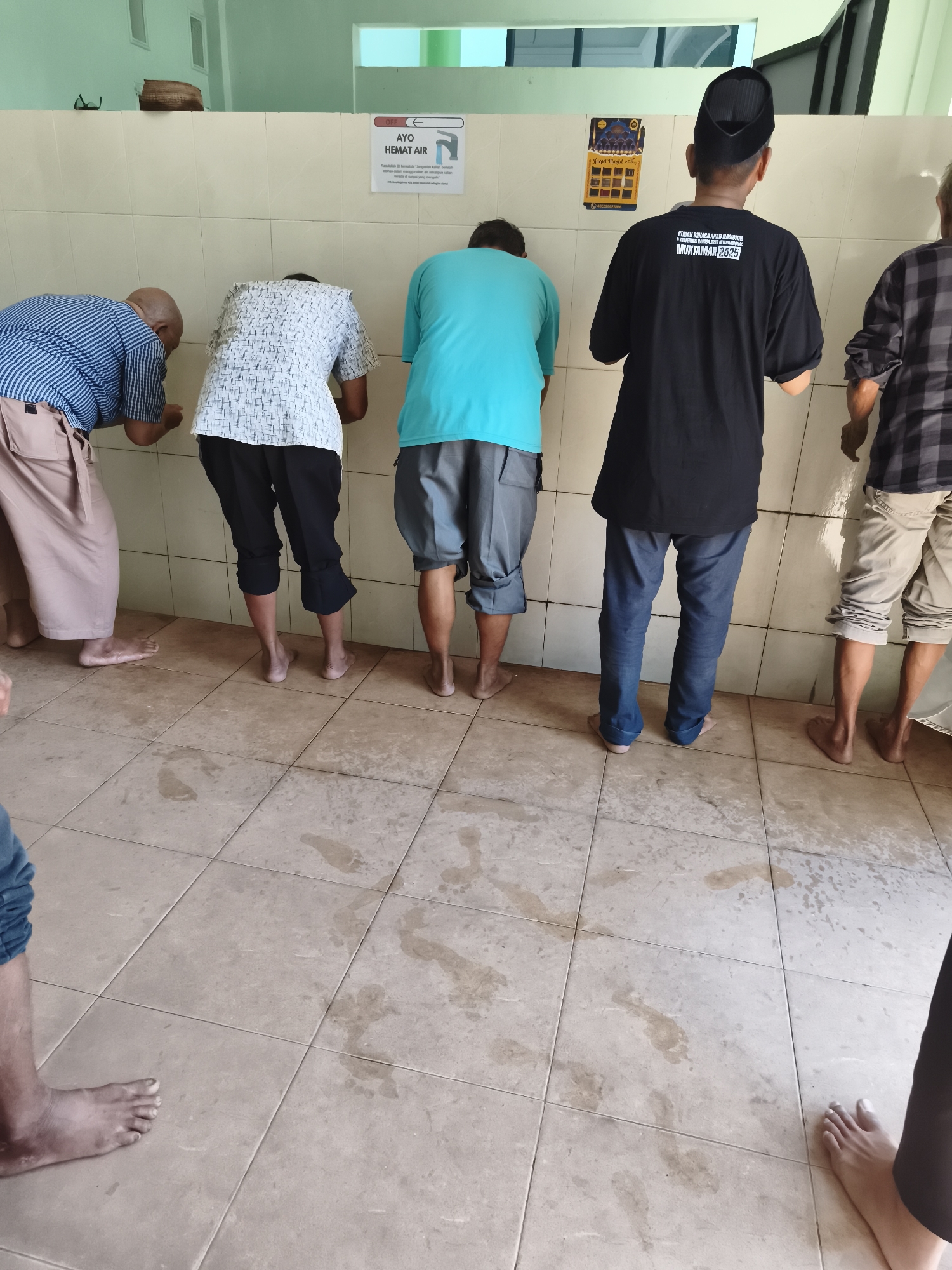 The image depicts a row of five men using a public urinal in a tiled room. The urinal is mounted on a beige tiled wall. The floor is also tiled with a light brown ceramic. Water stains are visible on the floor, suggesting recent use.
From left to right, the first man is bald and wears a blue and white checkered shirt with a light brown garment draped over his shoulders. He is leaning forward, using the urinal.
The second man wears a white shirt with a black and white geometric pattern and dark pants. He is also leaning forward, facing the urinal.
The third man wears a teal t-shirt and grey shorts. He is facing away from the camera, towards the urinal.
The fourth man wears a black t-shirt with white text and dark blue pants. He has a black peci (a traditional cap) on his head and is facing the urinal.
The fifth man, partially visible on the far right, wears a dark and light plaid shirt and light-colored shorts. He is also using the urinal.
Above the urinals, there is a sign with the text "AYO HEMAT AIR" (Let's Save Water) with an image of a tap. Another yellow poster with text and an image of a mosque is on the wall to the right of the first sign. The walls are tiled in a light beige color. The lighting in the room appears to be artificial, coming from ceiling lights which are not visible in the frame.
This scene is likely located in a public restroom facility, possibly in a mosque or a similar public building in Tonasa, Indonesia, given the cultural context suggested by the attire and the language on the sign. The presence of multiple men using urinals side-by-side indicates a communal restroom.