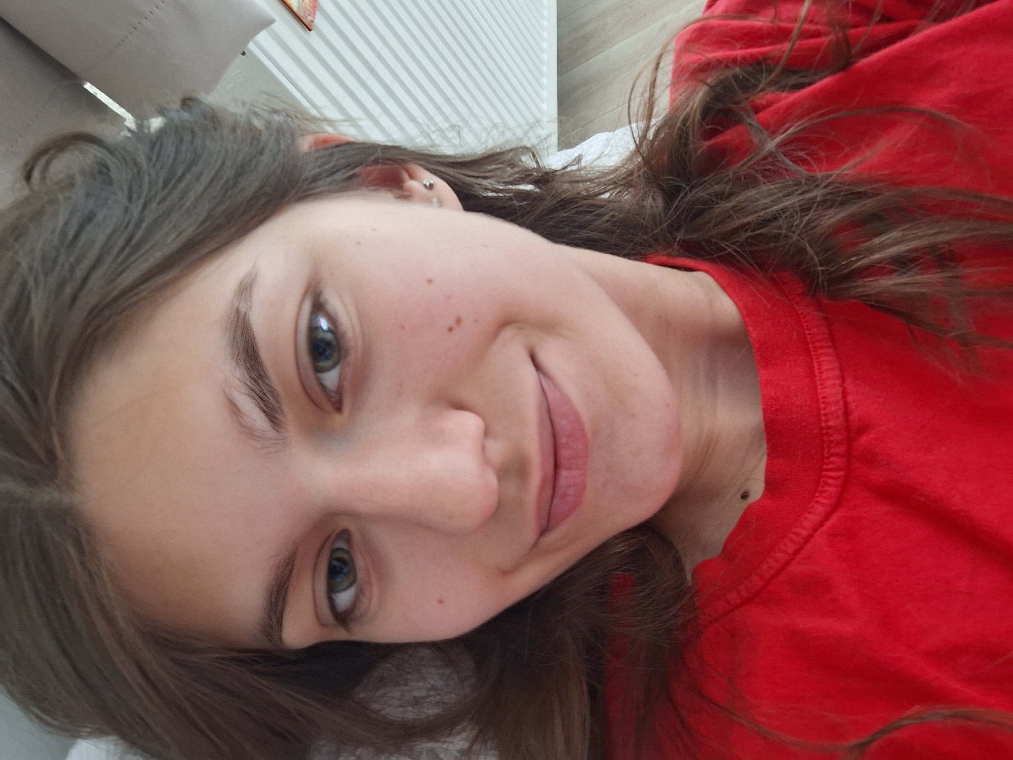 A young woman with light skin, brown hair, and green eyes is lying down. She is wearing a red shirt. The background includes a beige pillow and white blinds. The location in Panagyurishte, Bulgaria, cannot be determined from the image.