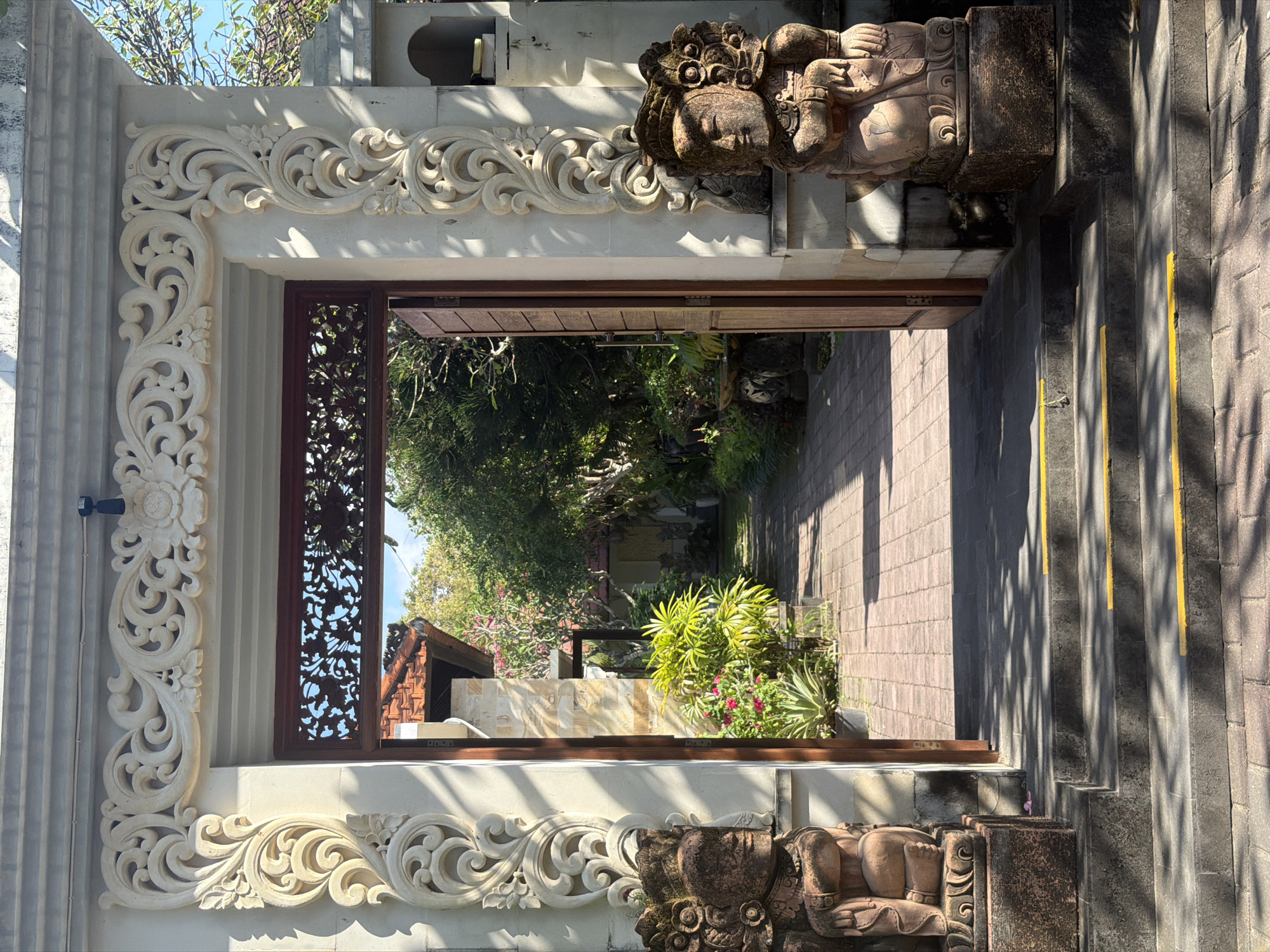 The image shows an ornate entrance to a building in Denpasar, Indonesia. Two stone statues of guardians, resembling Balinese mythological figures, stand on either side of the entrance. The entrance itself is framed by intricate white stonework carvings and a wooden gate that is partially open, revealing a walkway leading into a lush garden. The walkway is paved with red bricks, and tropical plants and trees are visible in the background. The sunlight casts shadows on the ground, indicating it is daytime. The architecture suggests a traditional Balinese style, common in temples or cultural sites within the city.