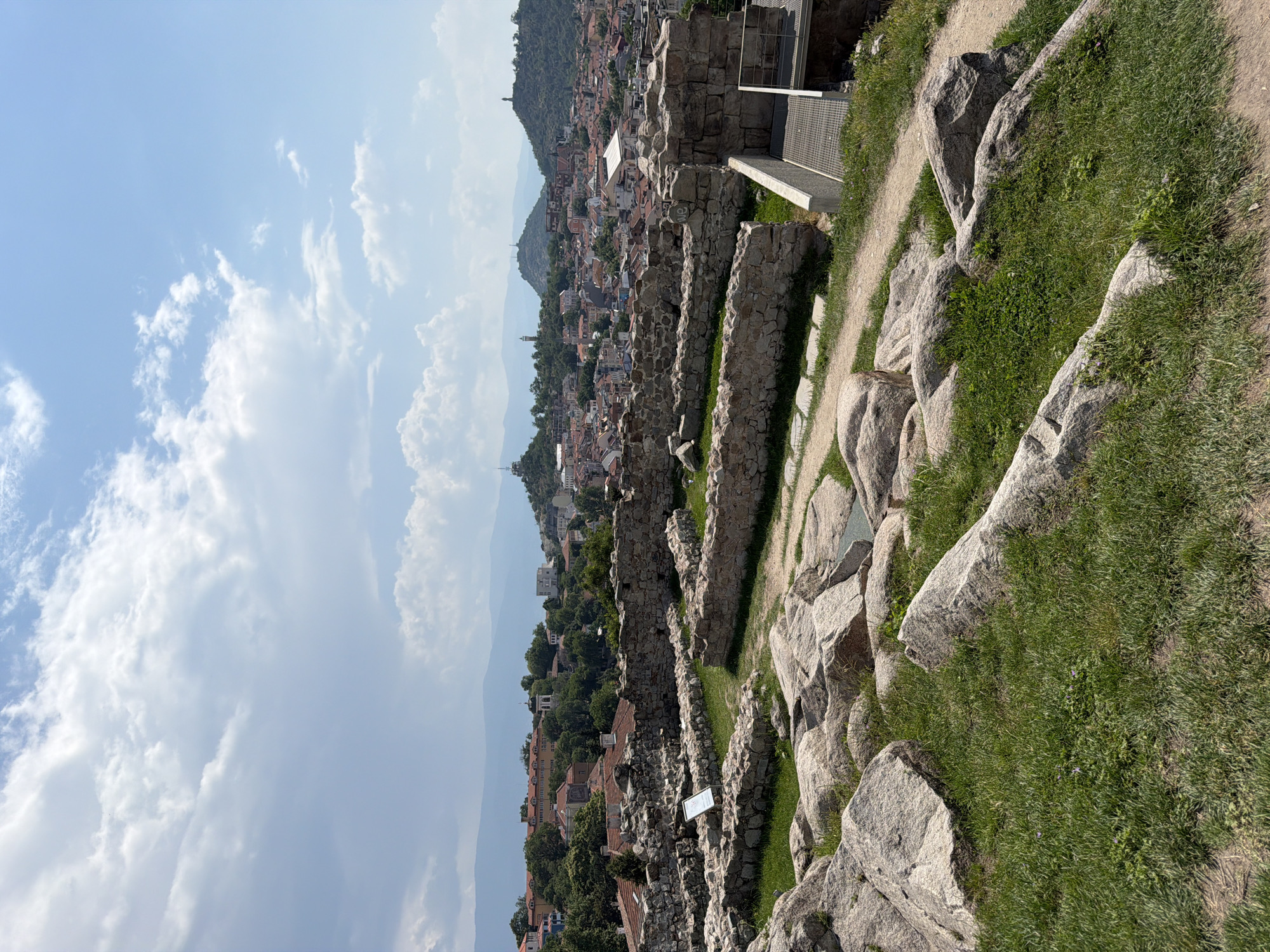An elevated, vertical shot captures an archaeological site in Plovdiv, Bulgaria, overlooking the city and distant mountains under a partly cloudy sky. The upper third of the image features a bright blue sky filled with scattered white cumulus clouds, denser on the left and more wispy on the right.
The foreground, occupying the lower third, consists of a grassy slope interspersed with numerous large, irregularly shaped grey boulders. A dirt path winds through this rocky terrain from the lower left, leading into the midground.
The midground showcases extensive archaeological ruins, characterized by low, rough-hewn stone walls and foundations forming rectangular enclosures. Green grass grows within these ancient structures. A white rectangular sign, possibly an informational plaque, stands erect near the ruins on the left side. On the far right, a modern-looking structure with a grey metal grate platform is visible, possibly providing access or serving as an observation point.
Beyond the ruins, the background reveals a panoramic view of the city of Plovdiv. A dense urban sprawl is visible, dominated by buildings with red-tiled roofs. Several prominent hills, or "tepes," rise from the cityscape; some are dotted with buildings and what appears to be a church spire on the left, while others feature distinct monuments or structures at their peaks, particularly on the central-right. In the furthest background, hazy blue mountain ranges stretch across the horizon.
The scene is illuminated by bright, natural sunlight, indicative of a clear day. No people or animals are present in the image.
