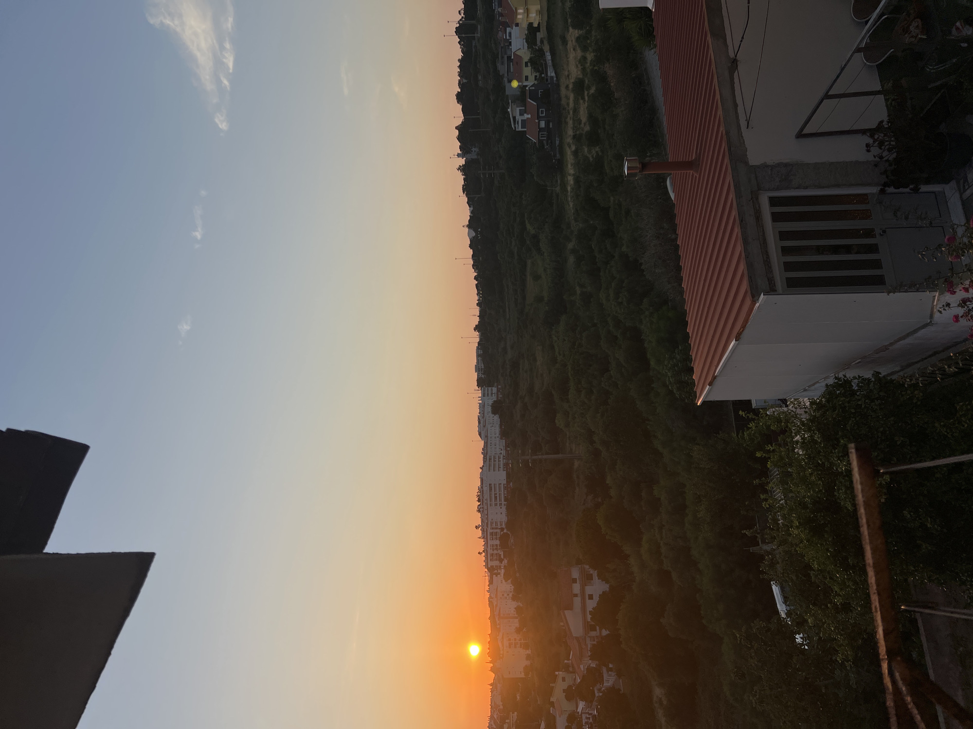 The image captures a sunset over a suburban landscape in Cascais, Portugal. The sun, a bright orange orb, is low on the horizon, casting a warm glow across the sky and illuminating a cluster of buildings in the distance. The foreground features a rooftop structure with a red corrugated metal roof and a white wall, a chimney, and a grey door with vertical glass panes. A railing and dense green foliage are also visible. Beyond this, the landscape unfolds with rolling hills covered in a thick canopy of trees and vegetation. Scattered among the trees are residential buildings with red roofs. The sky is a gradient of pale orange near the horizon, transitioning to a soft blue, with a few wispy clouds. The overall scene suggests a peaceful evening in a coastal Portuguese town.