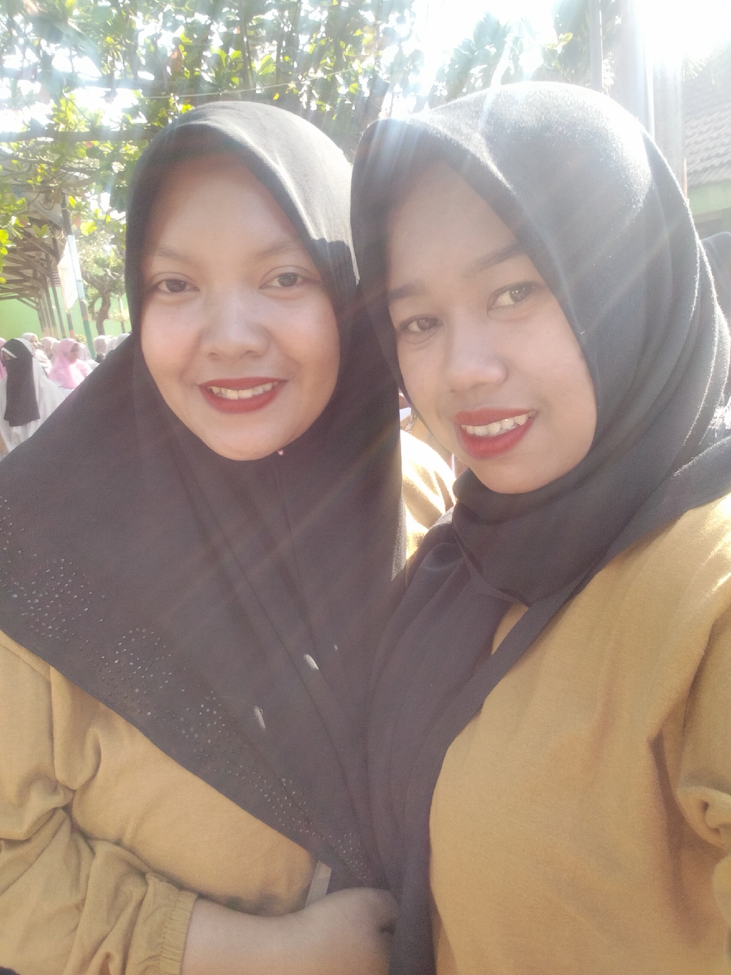 The image shows two women wearing hijabs and yellow tops. The woman on the left has a black hijab with rhinestones. She is smiling and wearing red lipstick. The woman on the right is also smiling and wearing red lipstick. They appear to be outdoors, possibly in a schoolyard or public area, with trees and buildings visible in the background. There are other people in the background wearing pink and white hijabs. The overall lighting suggests a sunny day. The scene appears to be a casual selfie taken by the two women. Based on the given location of Puspo, Indonesia, this image likely depicts a scene within a community or educational setting in that area.