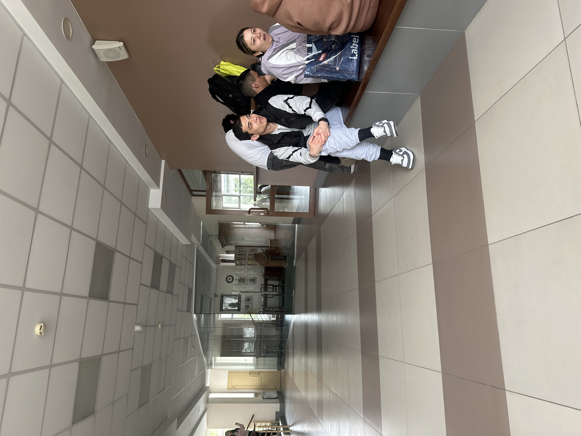 Four young people are seated on a wooden bench in an indoor hallway, likely in a public building. In the foreground, a man with dark hair, wearing a white t-shirt, a black and white vest, light gray sweatpants, and white sneakers with black socks, is seated with his hands clasped, looking towards the camera with a slight smile. Beside him, a woman with dark hair, dressed in a light purple top, looks towards the camera with a gentle expression. A blue bag with "label" printed on it rests next to her. Partially visible behind them are another person in a white t-shirt and a person in a light brown hoodie. Yellow and black backpacks are also near the group.
The hallway features a tiled floor with alternating light beige and brown stripes, creating a strong linear perspective. The ceiling is made of white panels with visible grid lines, equipped with recessed lighting and a white speaker-like device. On the right side of the hallway, glass partitions reveal an interior space containing furniture, framed pictures, and a clock. Mirrored on the glass, the Cyrillic text "МУЗЕЙ НЕВРОЛОГИИ" is visible, translating to "MUSEUM OF NEUROLOGY," indicating the location. Further down the hall, a light-colored door is seen, and a person is partially visible in the distant background. The scene depicts a casual daytime moment, with the individuals appearing relaxed, possibly waiting or socializing.