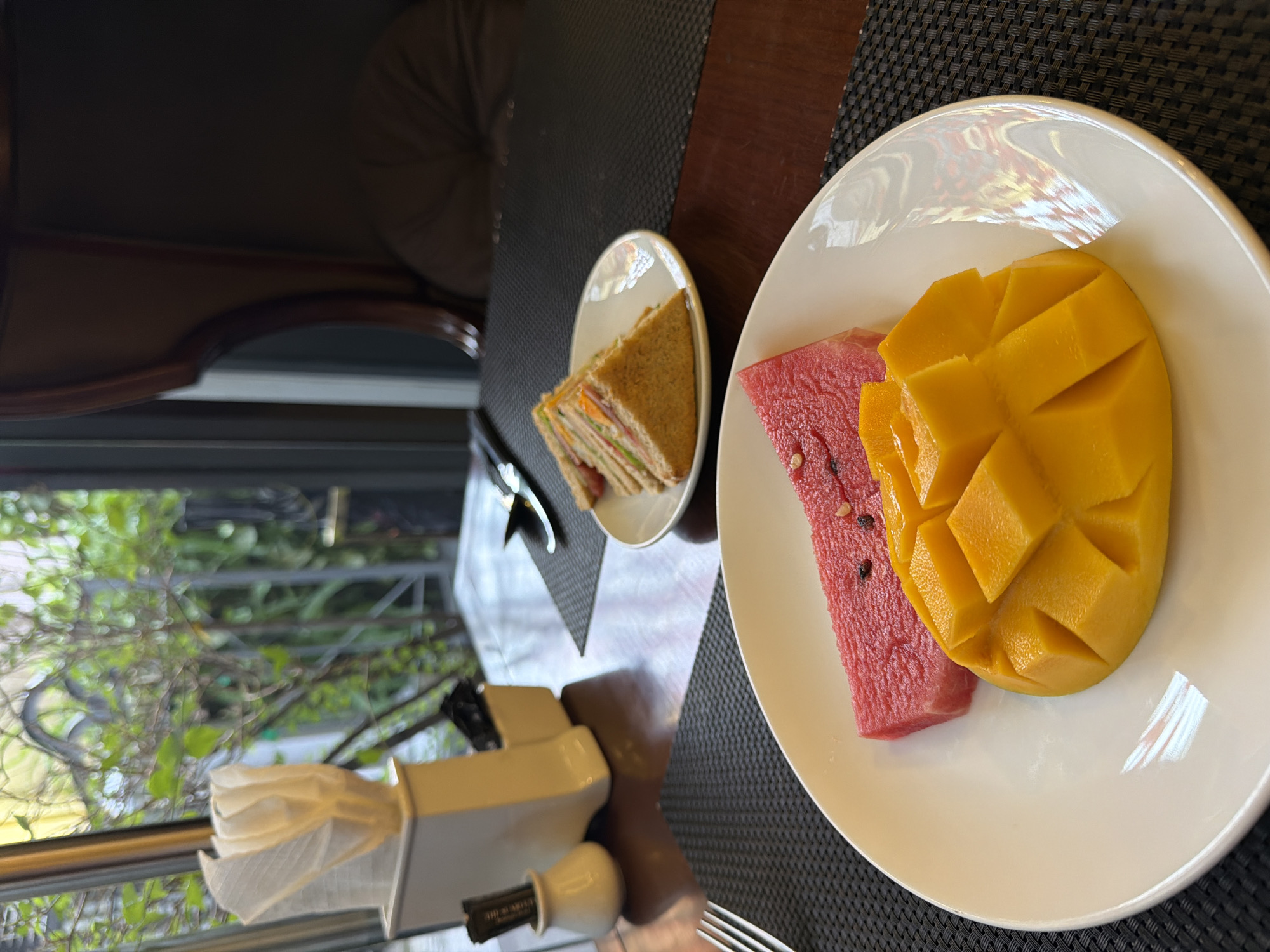 A plate of fruit, consisting of sliced watermelon and a diced mango, is positioned in the foreground on a dark placemat. To the left, a fork rests on the placemat. In the background, a white ceramic holder containing napkins and sugar packets sits on a wooden table. Further back, a small plate holds a triangular sandwich, and a knife and fork are visible on a placemat. Beyond the table, a window reveals a view of green foliage. A plush, dark armchair is partially visible on the right side of the image. The scene appears to be set in a dining area, possibly within a hotel or restaurant in Thuận Hóa Ward, Vietnam, given the context provided.
